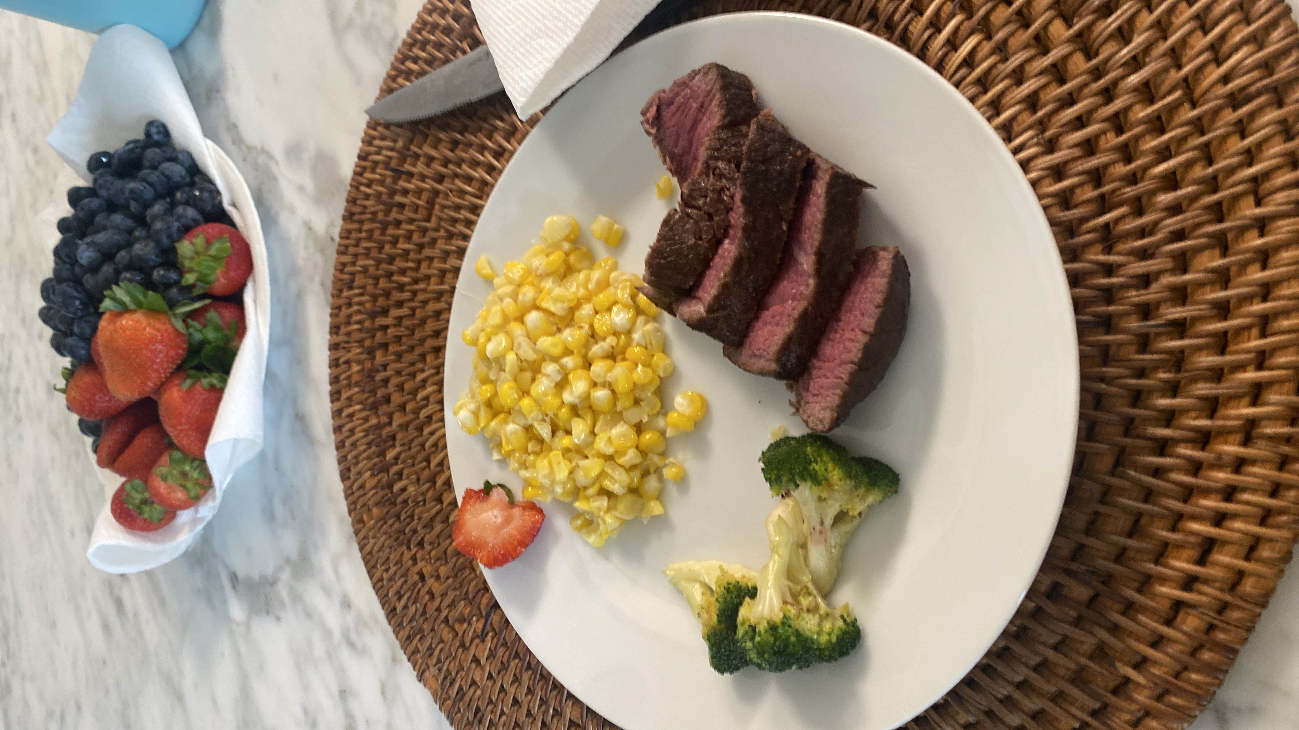 A balanced meal consisting of steak, corn, broccoli, and a side of fresh strawberries and blueberries.