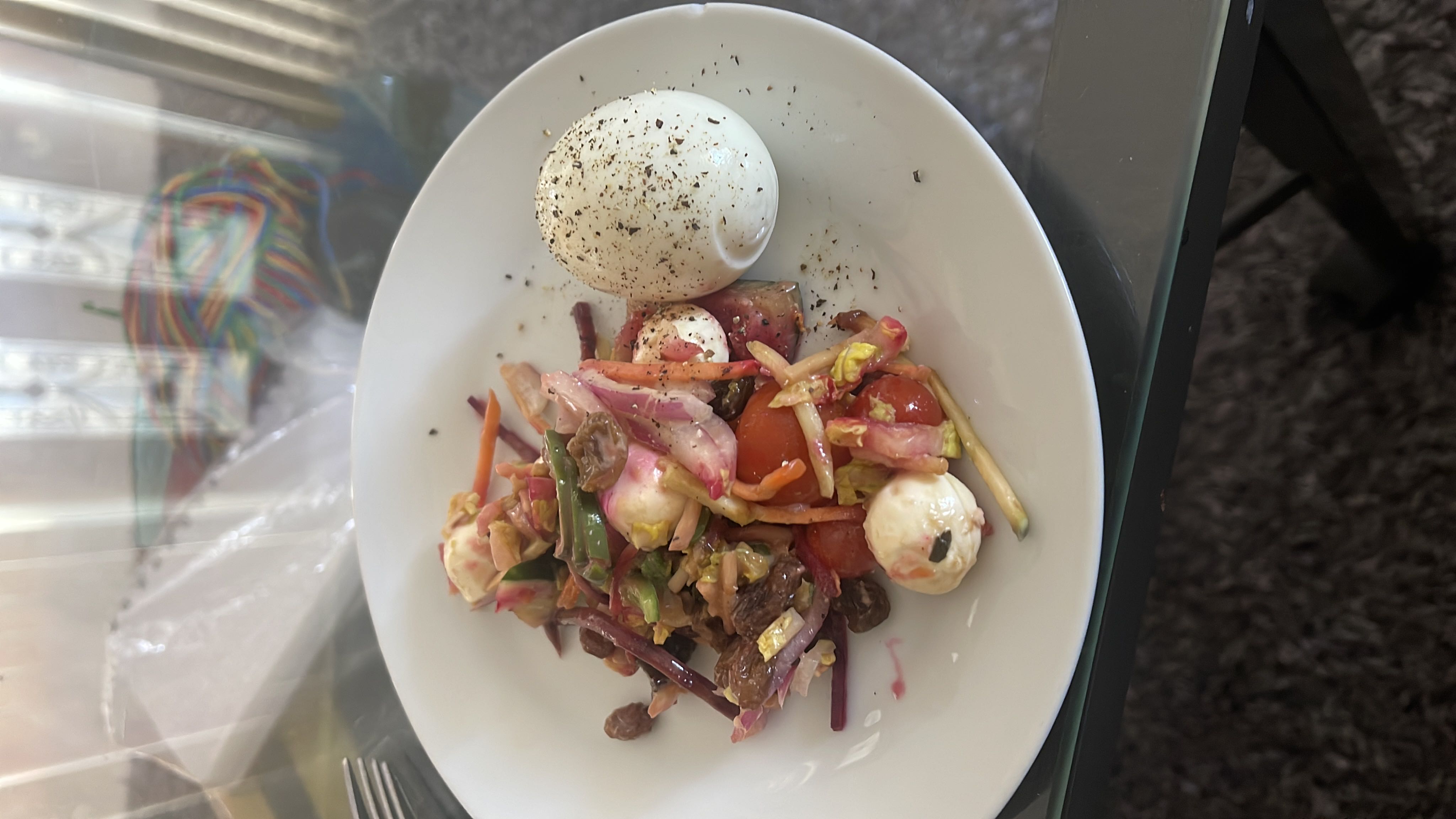 A plate containing a boiled egg and a mixed salad with vegetables, mozzarella, and raisins, seasoned with spices.