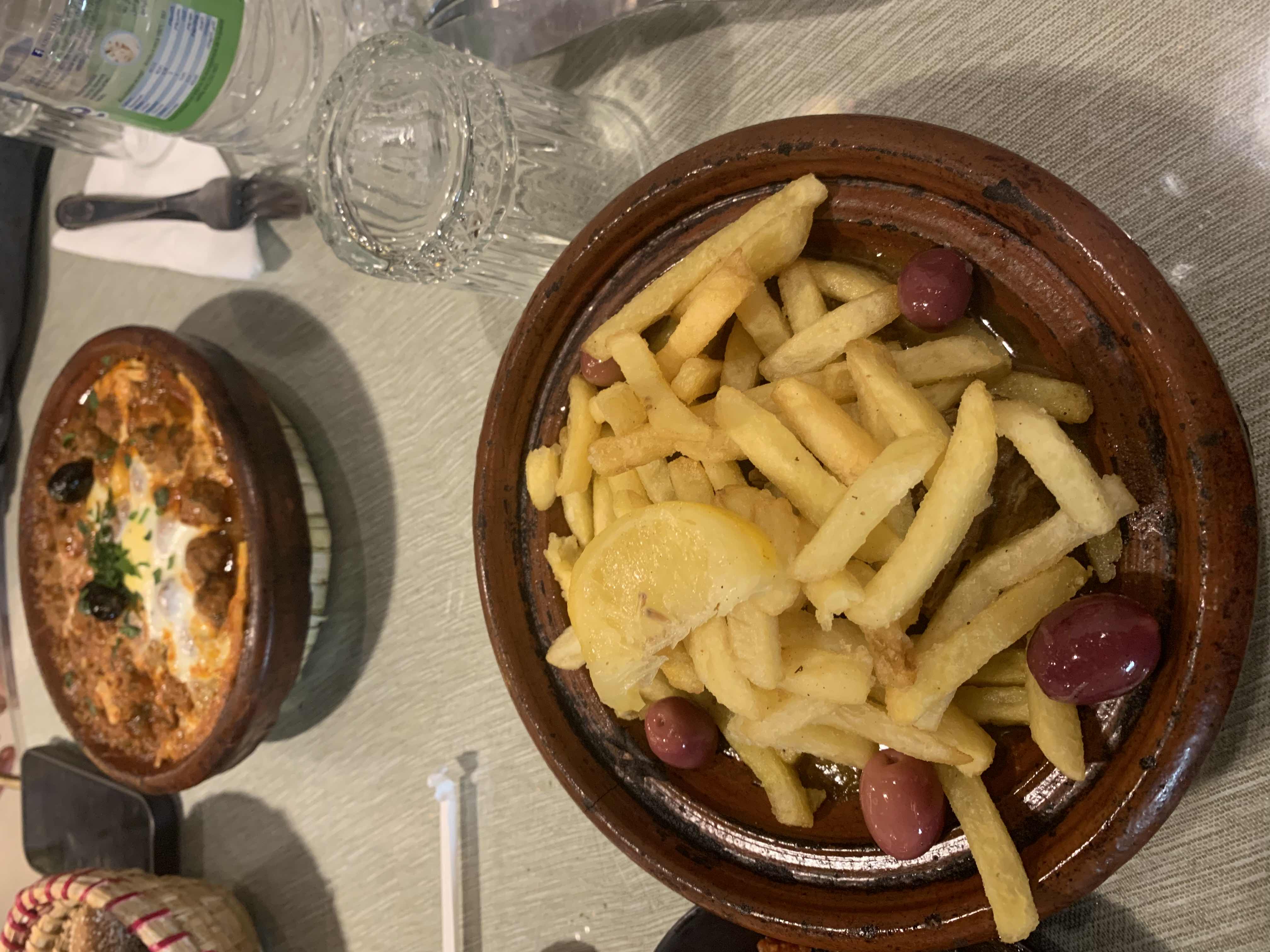 The image shows a dish of French fries with olives and a lemon slice, accompanied by a meat stew in a clay pot.