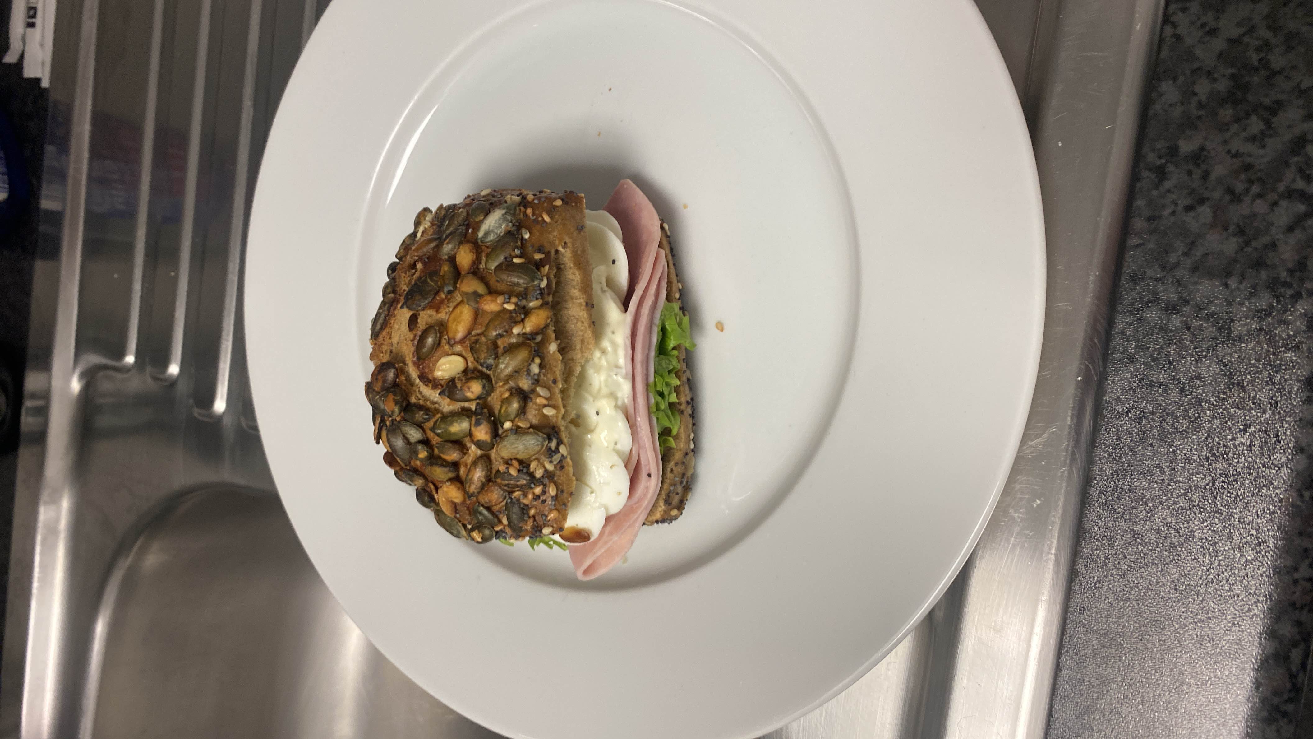 A sandwich made with a seeded bread roll, ham, lettuce, cheese, and mayonnaise, offering a balanced mix of protein, carbs, and fats.