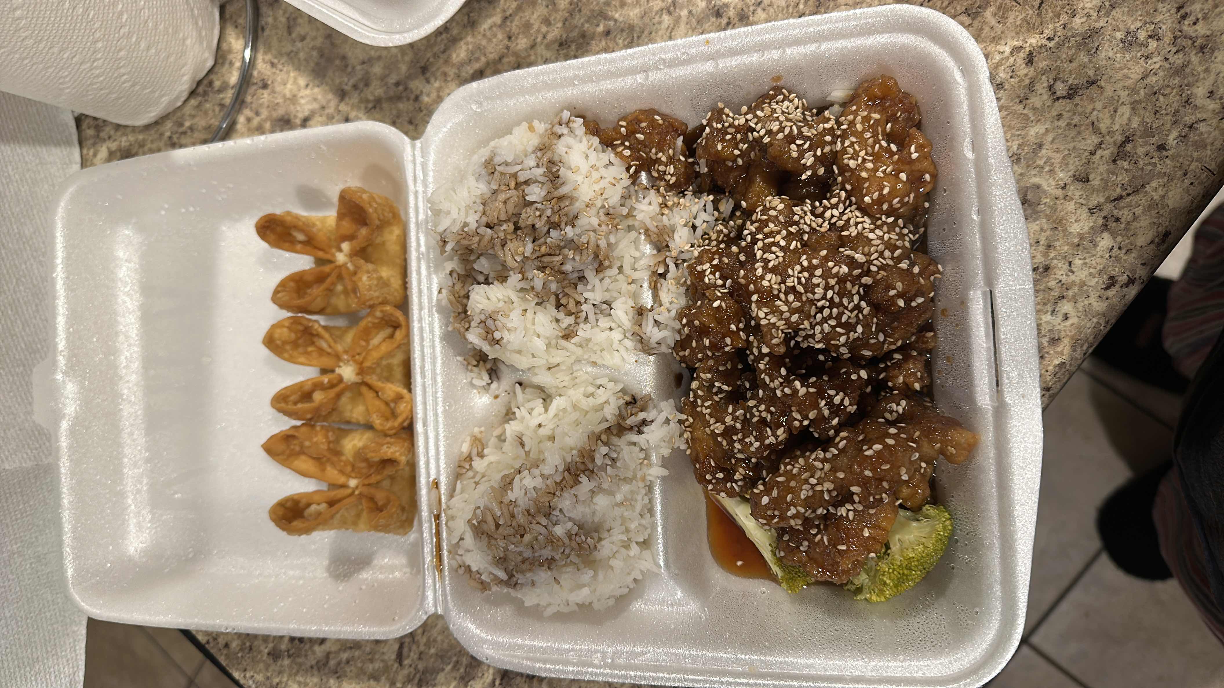 A takeout meal consisting of sesame chicken topped with sesame seeds, white rice, and fried wontons.