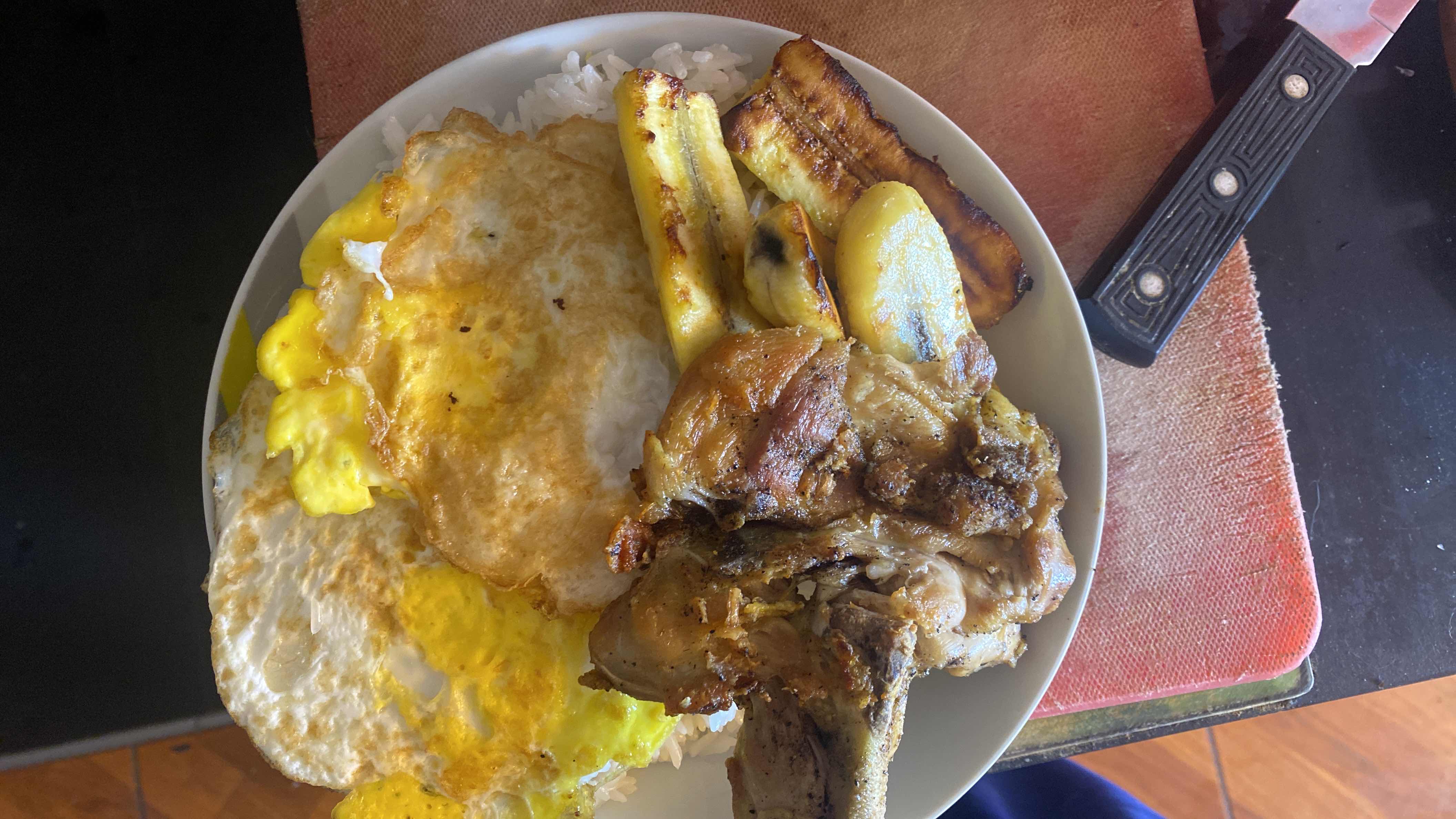A plate containing fried eggs, grilled chicken, white rice, and fried plantains, providing a balanced mix of protein, carbs, and fats.