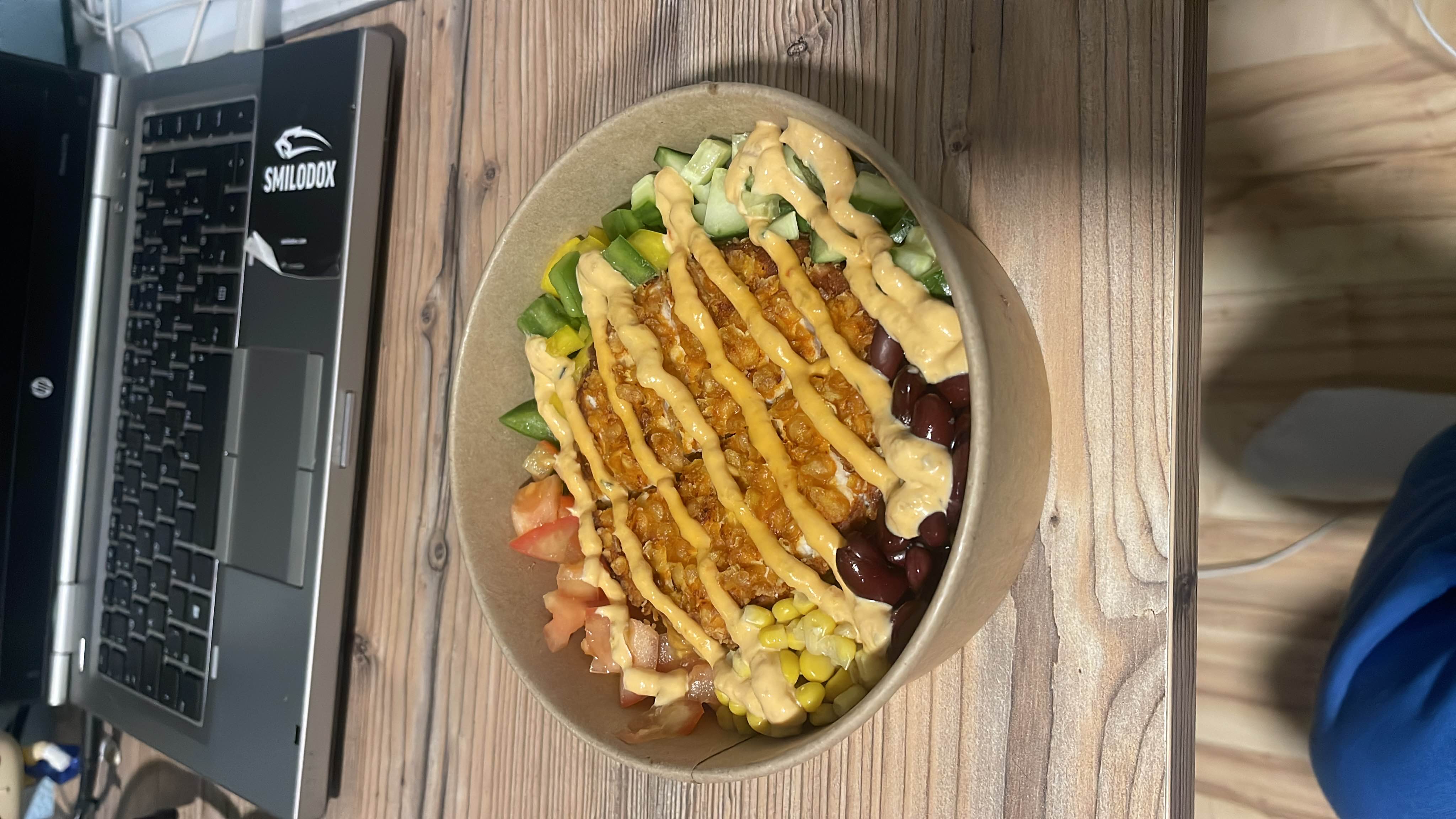 A colorful salad bowl featuring a crispy patty, beans, corn, fresh vegetables, and a creamy sauce.