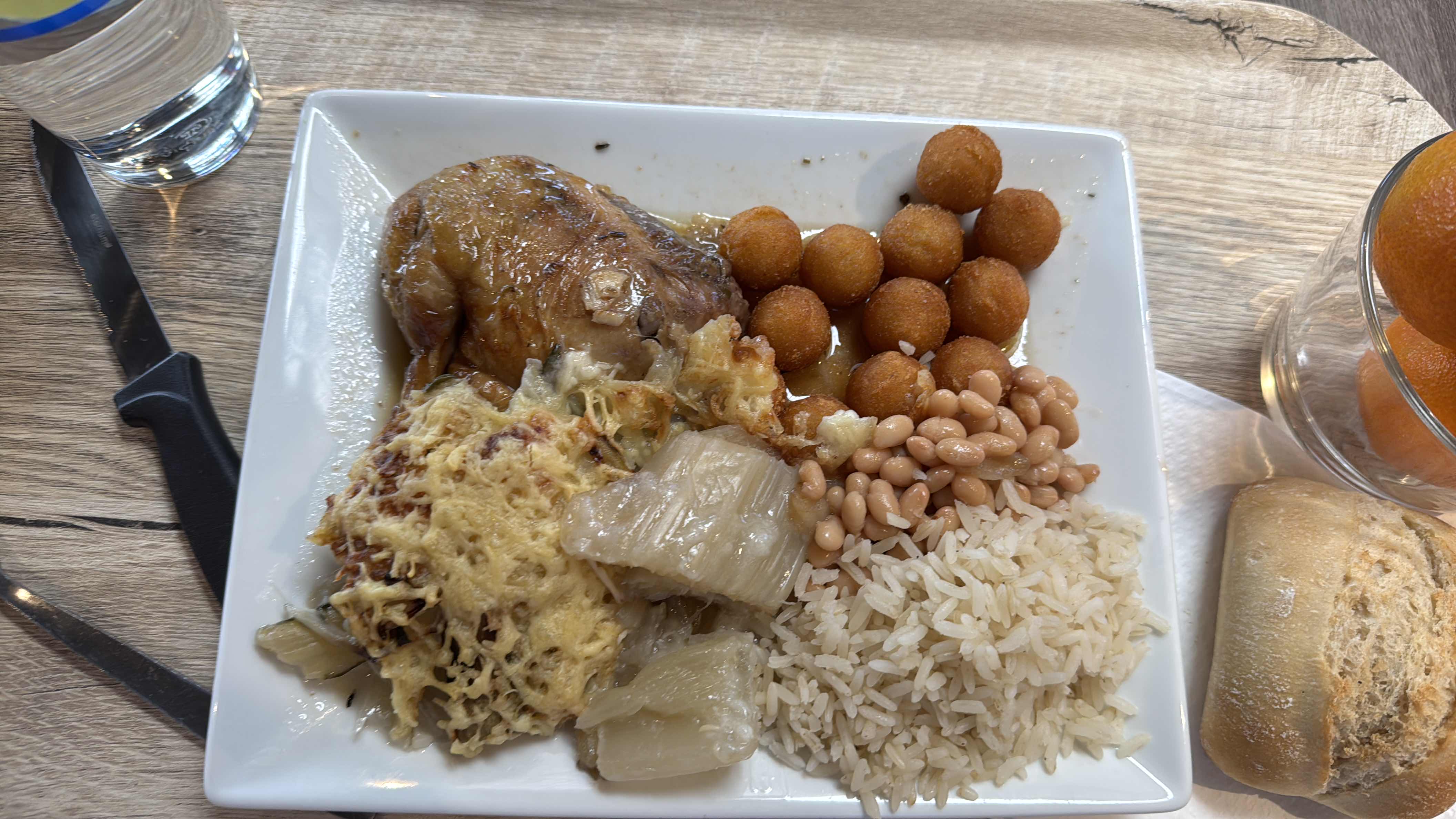 A balanced meal consisting of a chicken leg, rice, beans, fried balls, cabbage gratin, and a bread roll, providing a mix of protein, carbs, and fats.
