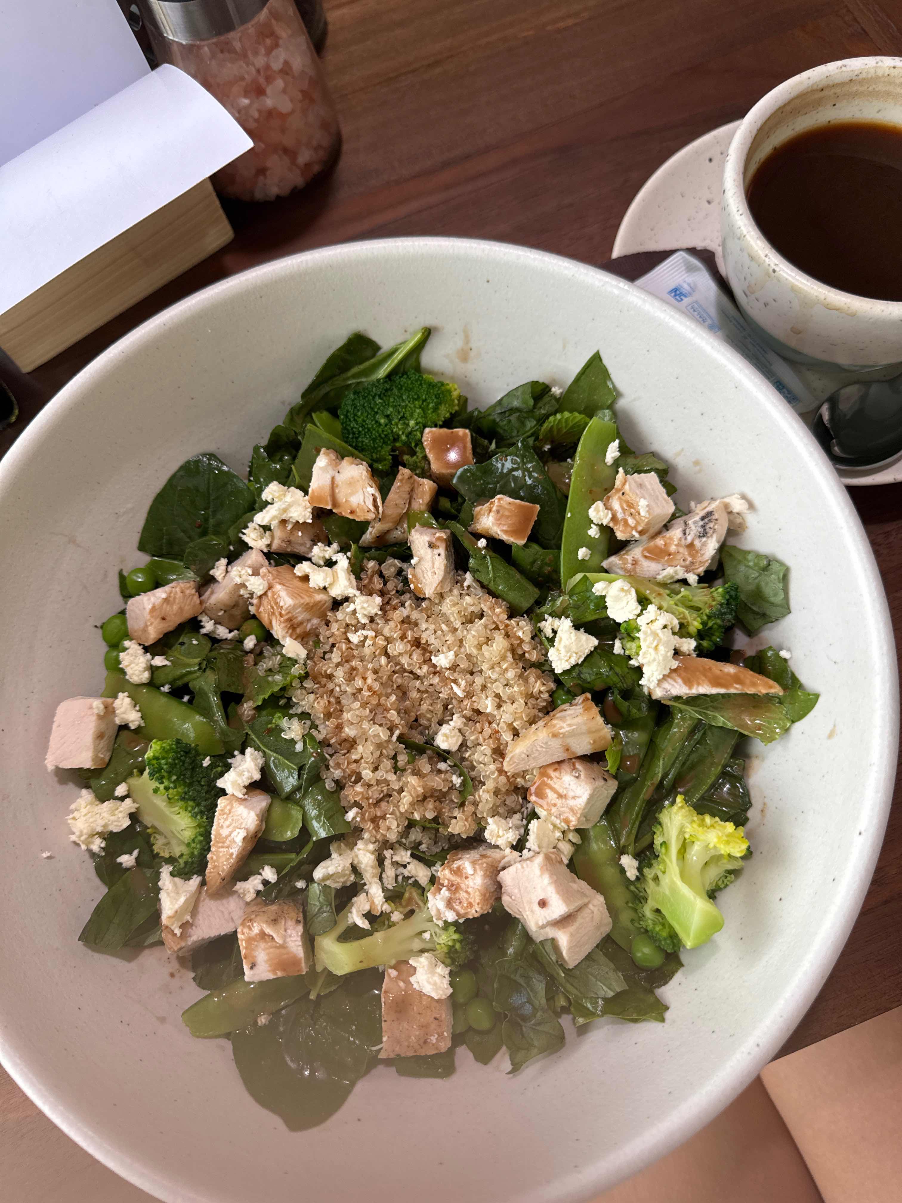 A healthy salad bowl containing chicken, quinoa, broccoli, spinach, and feta cheese, served with a cup of black coffee.