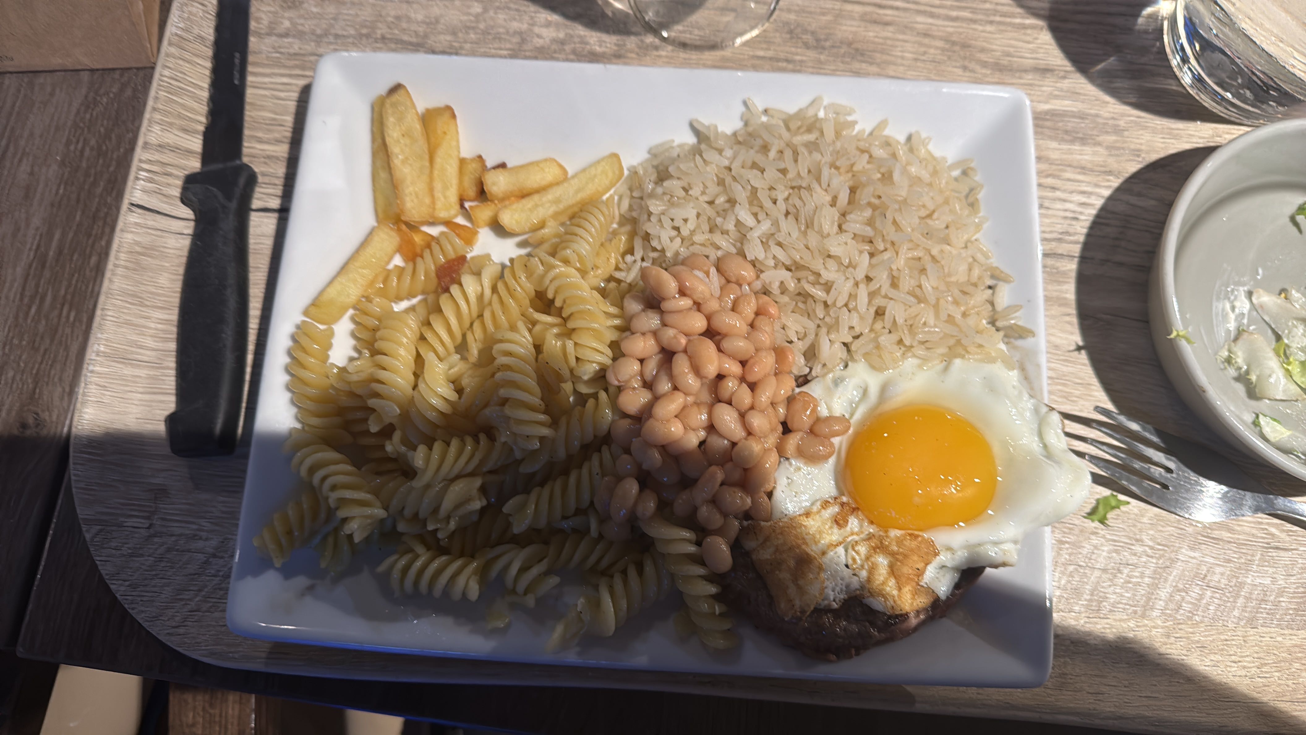 A mixed plate containing pasta, rice, fried egg, baked beans, and french fries, providing a balanced mix of carbs, protein, and fats.