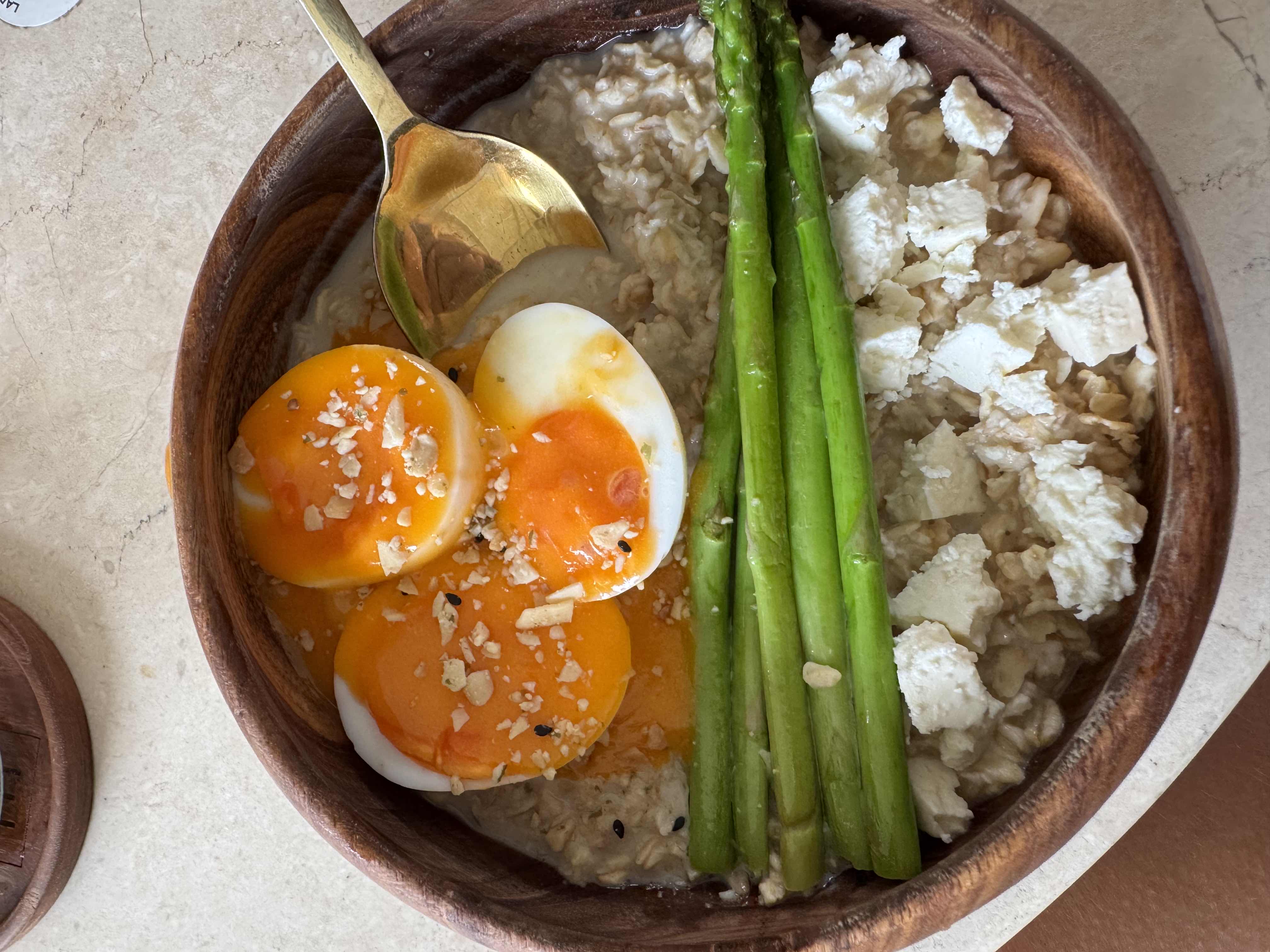 A nutritious bowl containing soft-boiled eggs, asparagus, oats, and crumbled feta cheese.