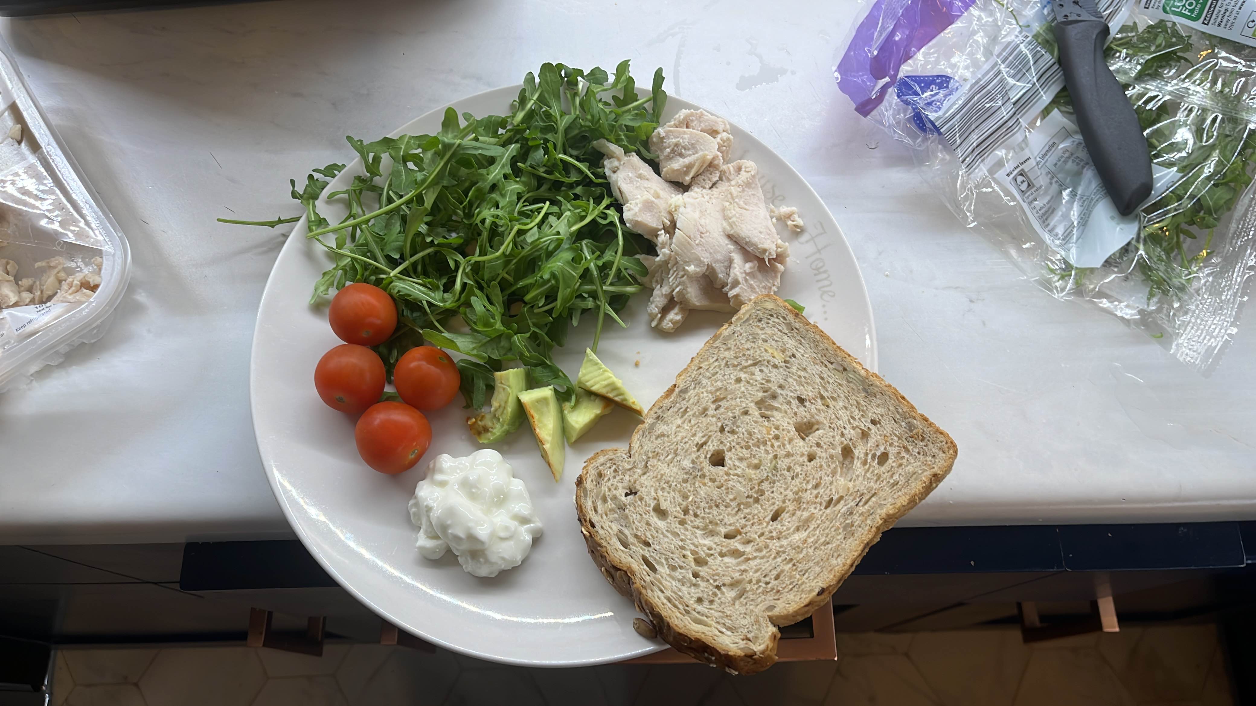 A balanced meal consisting of whole grain bread, chicken breast, arugula, cherry tomatoes, avocado, and cottage cheese, providing a good mix of protein, carbs, and healthy fats.