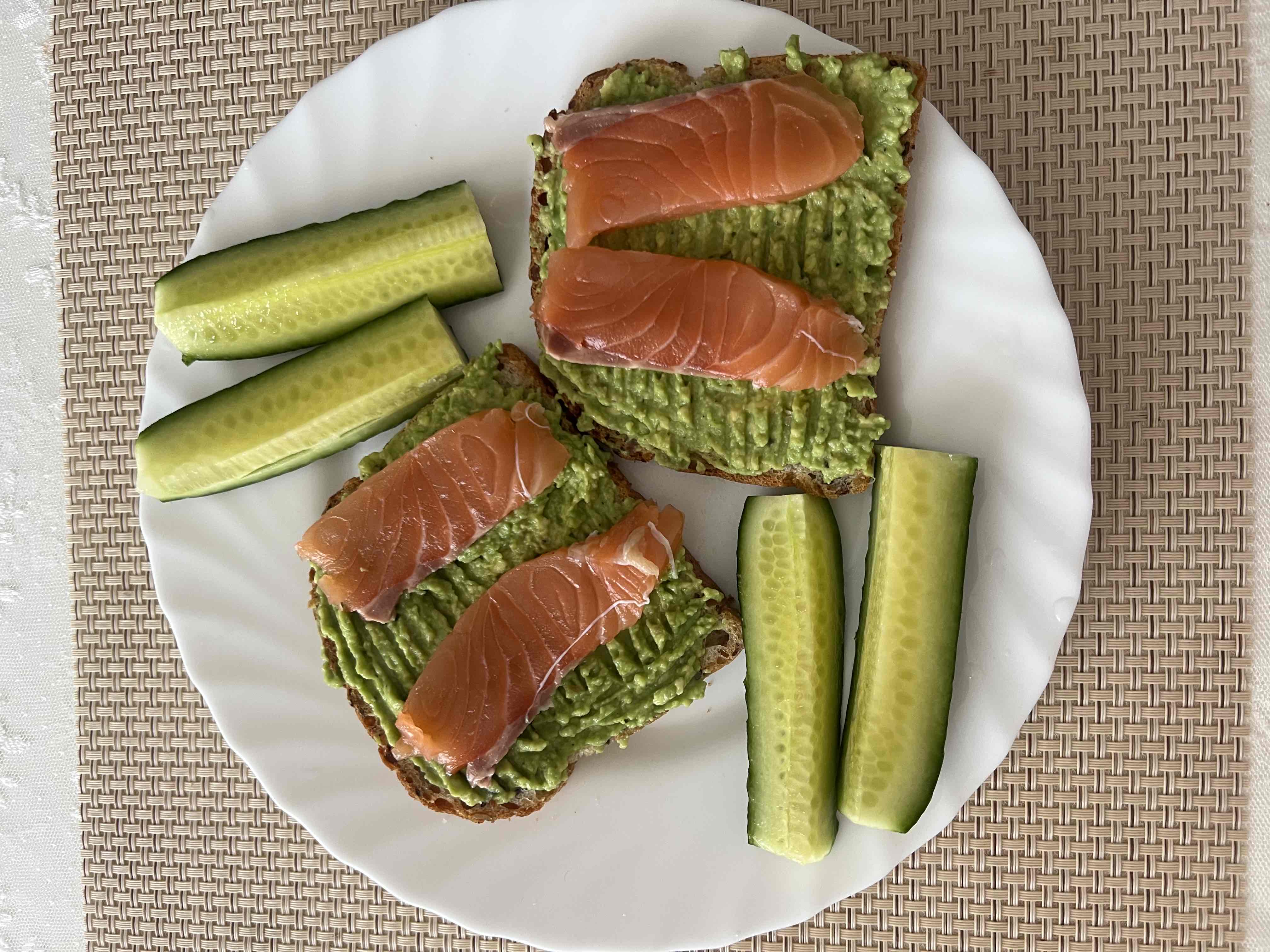 This dish consists of avocado spread on bread, topped with salmon slices, and served with cucumber sticks. It is a healthy and balanced meal.