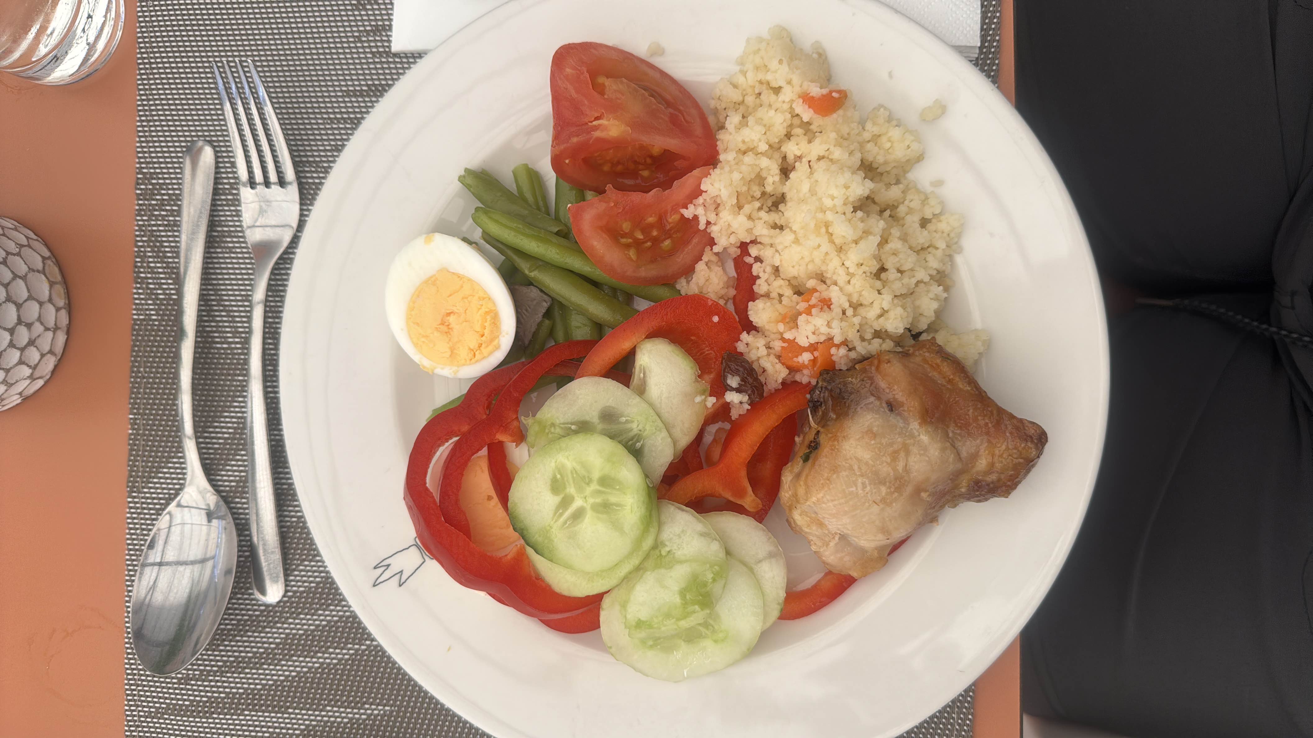 A balanced meal consisting of chicken, couscous, boiled egg, and fresh vegetables including cucumber, tomato, red bell pepper, and green beans.