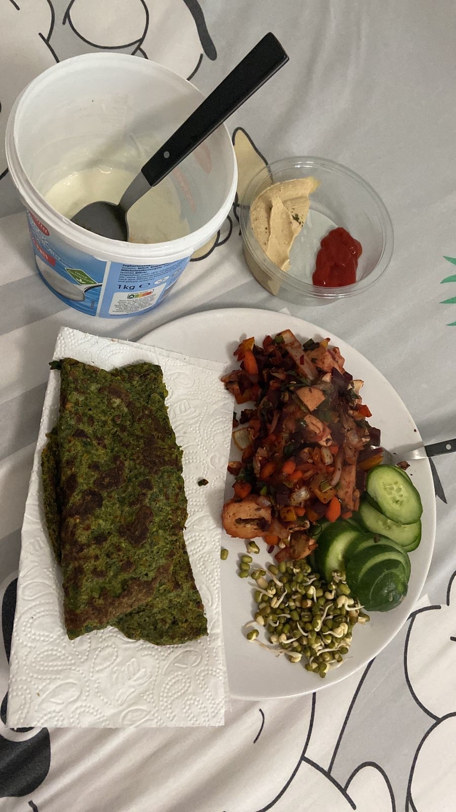 A balanced meal consisting of spinach pancakes, sauteed vegetables, cucumber slices, sprouts, hummus, ketchup, and yogurt, providing a mix of protein, carbs, and fats.