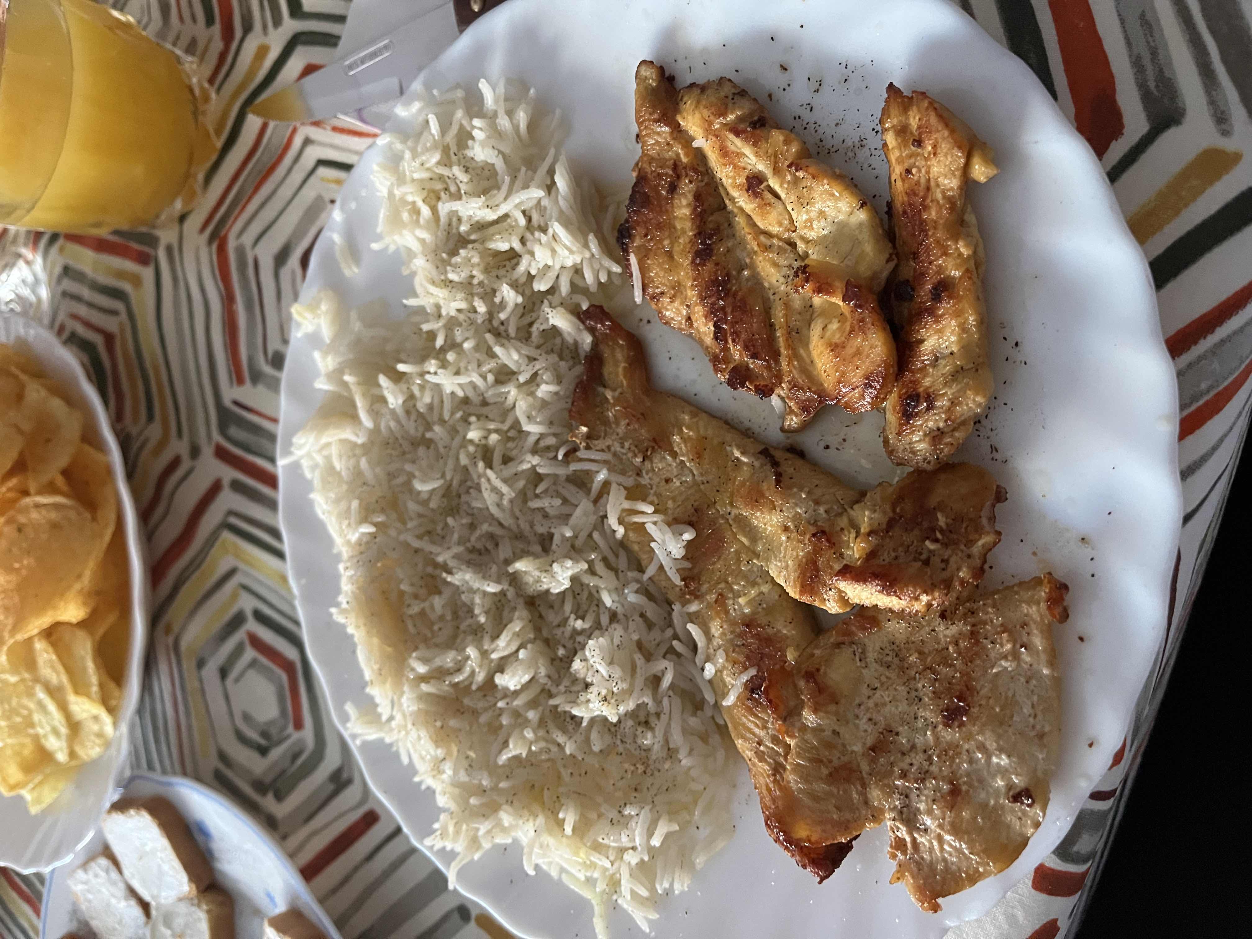 A meal consisting of grilled chicken, white rice, potato chips, and orange juice, providing a balanced mix of protein, carbs, and fats.