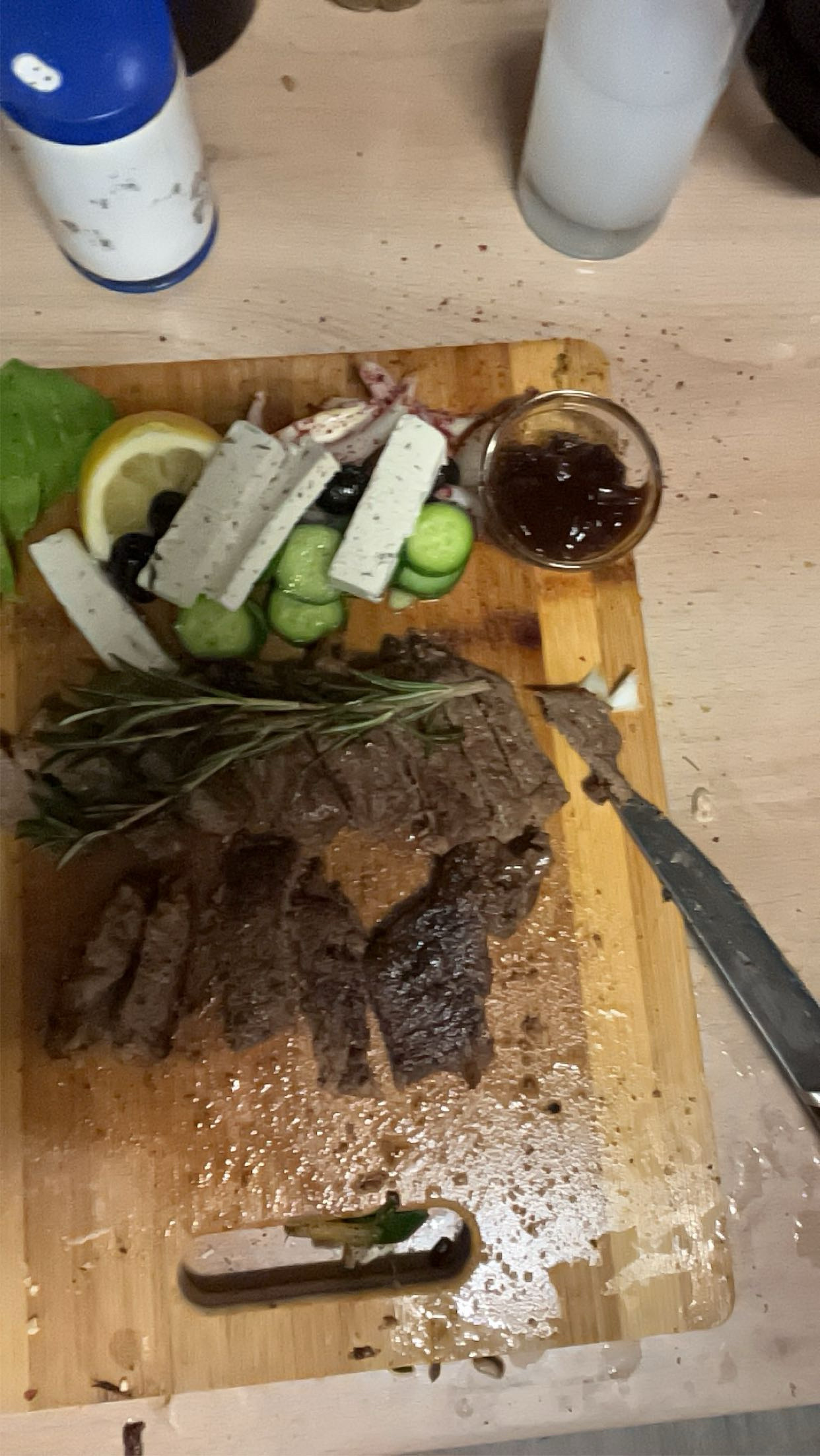 A meal consisting of grilled steak, feta cheese, cucumber slices, black olives, and a small portion of jam or sauce, served on a wooden board.