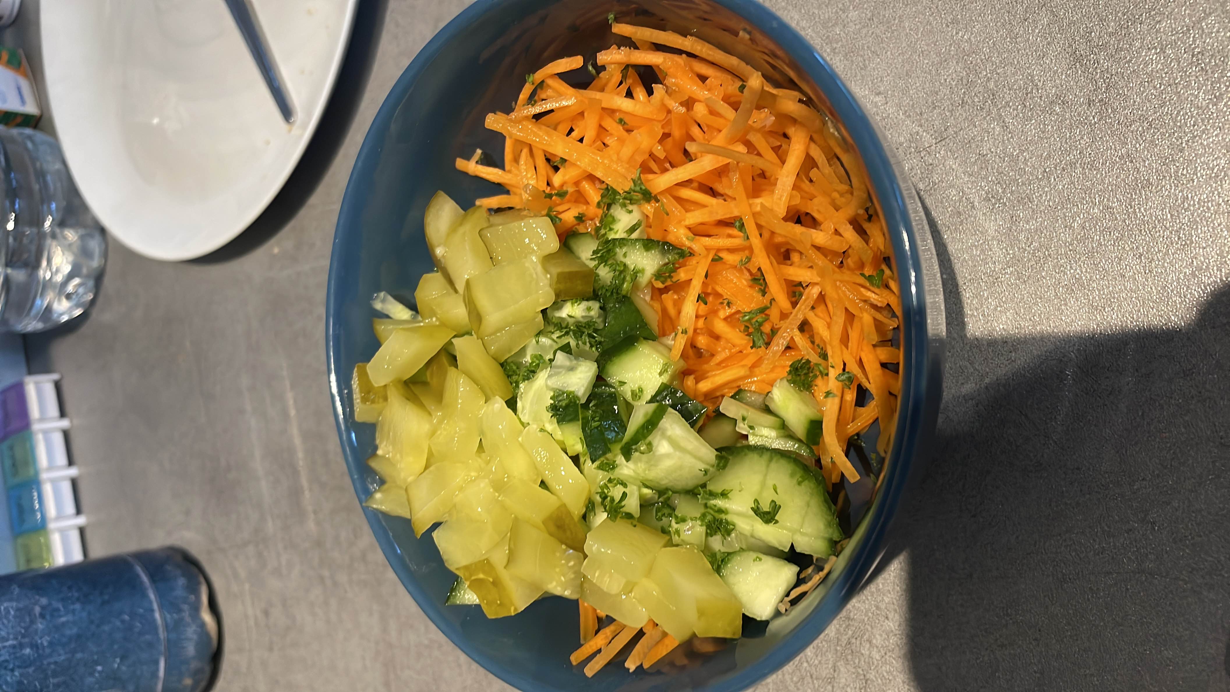 A fresh vegetable salad containing shredded carrots, sliced cucumbers, and chopped pickles, garnished with parsley.