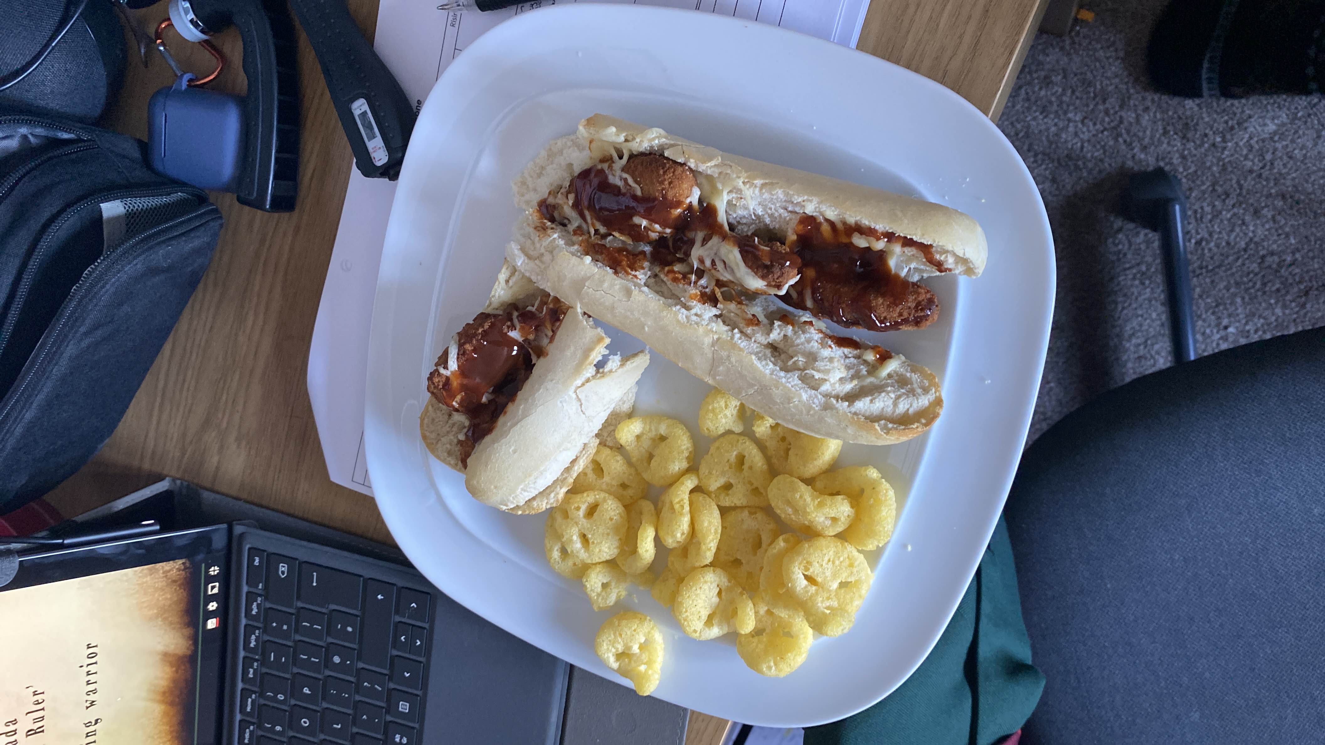 A meal consisting of two hot dog buns filled with chicken tenders, barbecue sauce, and cheese, served with a side of potato snacks.