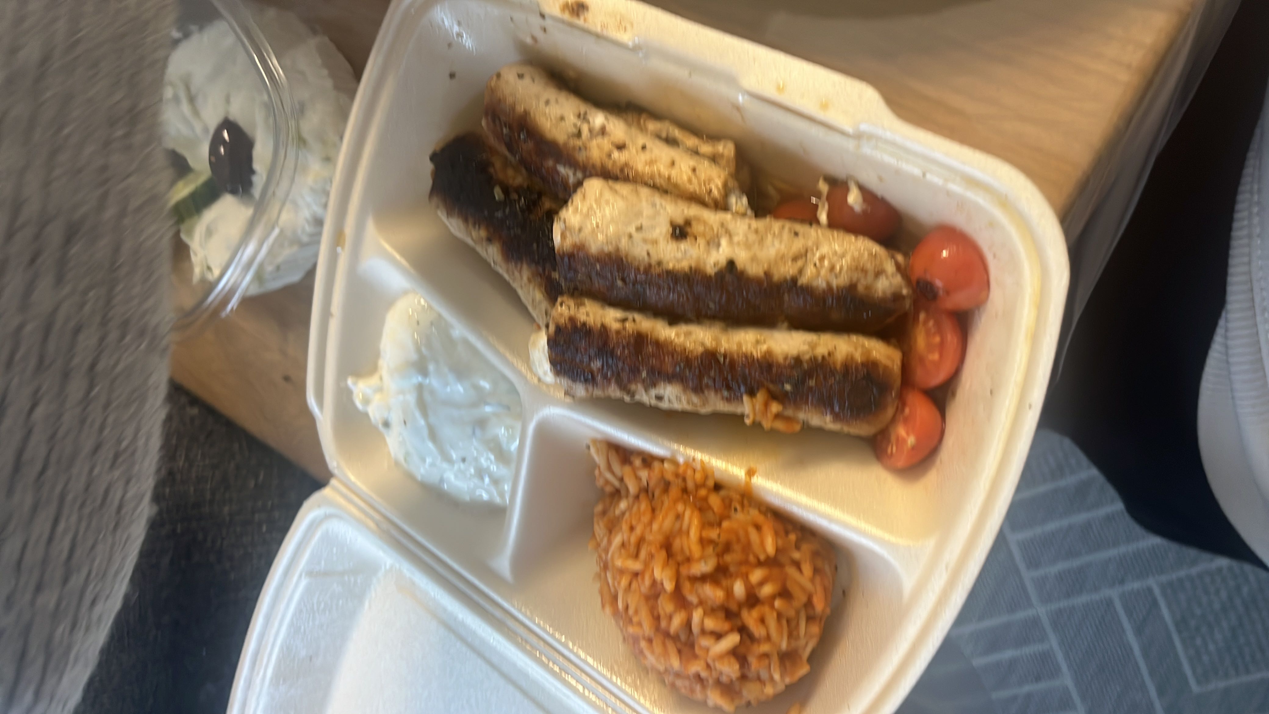 A meal consisting of grilled sausages, cherry tomatoes, rice, and tzatziki sauce, offering a balanced mix of protein, carbs, and fats.