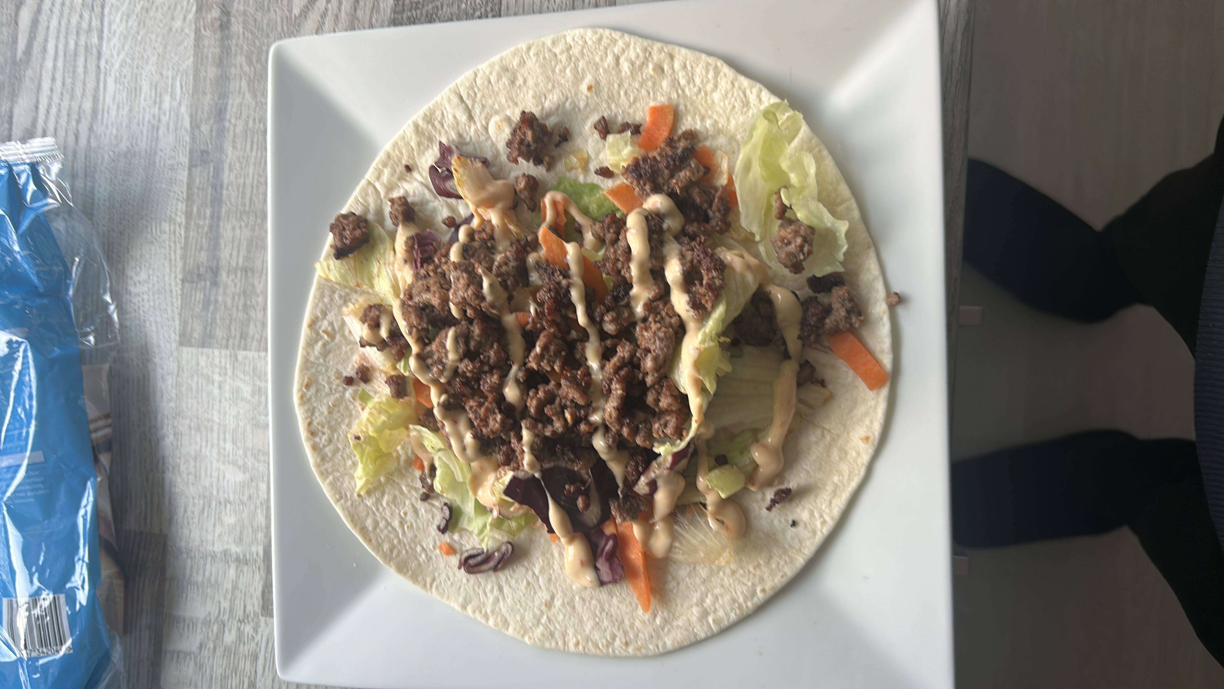 A tortilla wrap filled with ground beef, lettuce, carrots, and a drizzle of sauce, providing a balanced mix of protein, carbs, and fats.