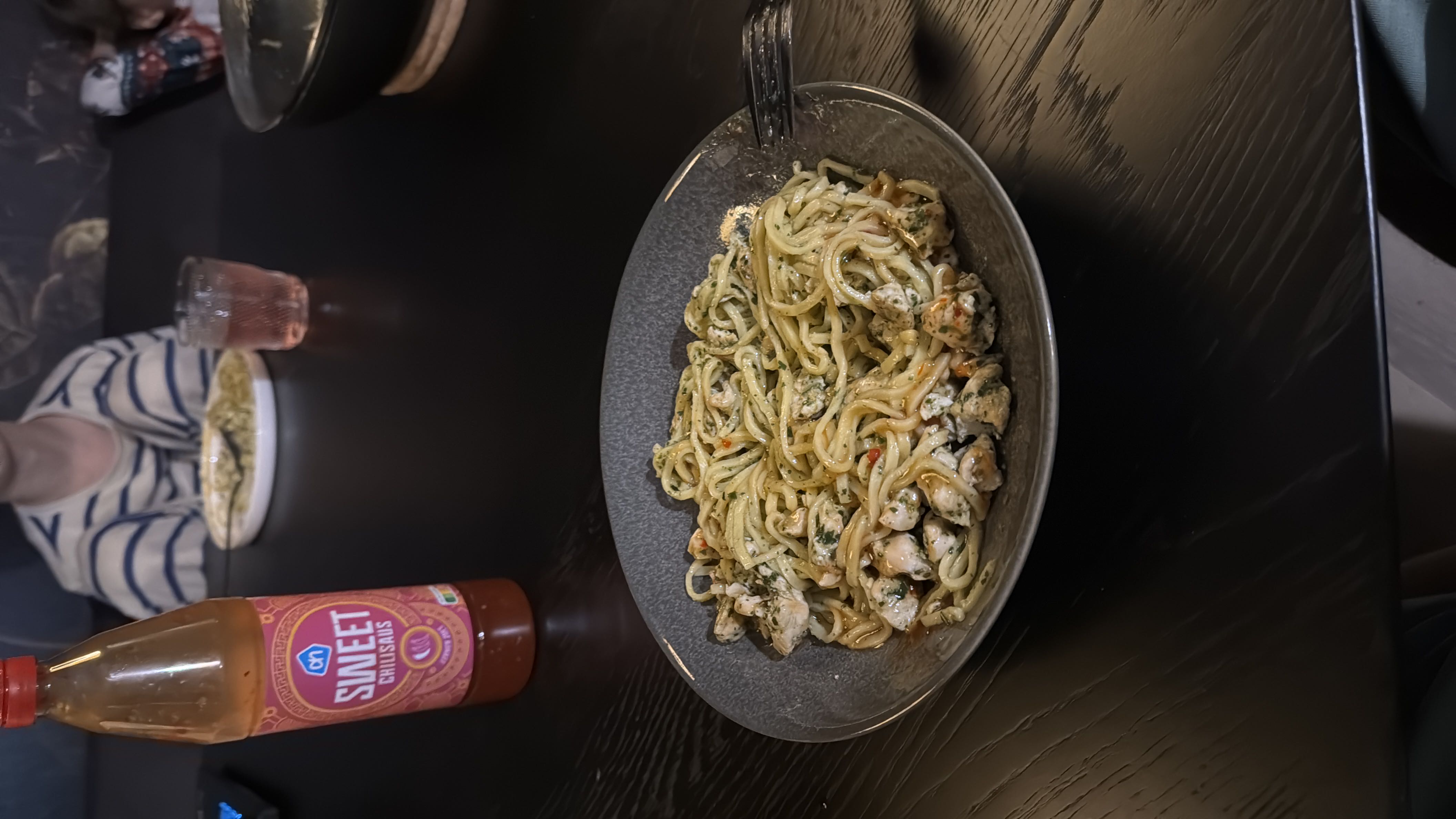 A plate of noodles with chicken and pesto sauce, accompanied by sweet chili sauce.