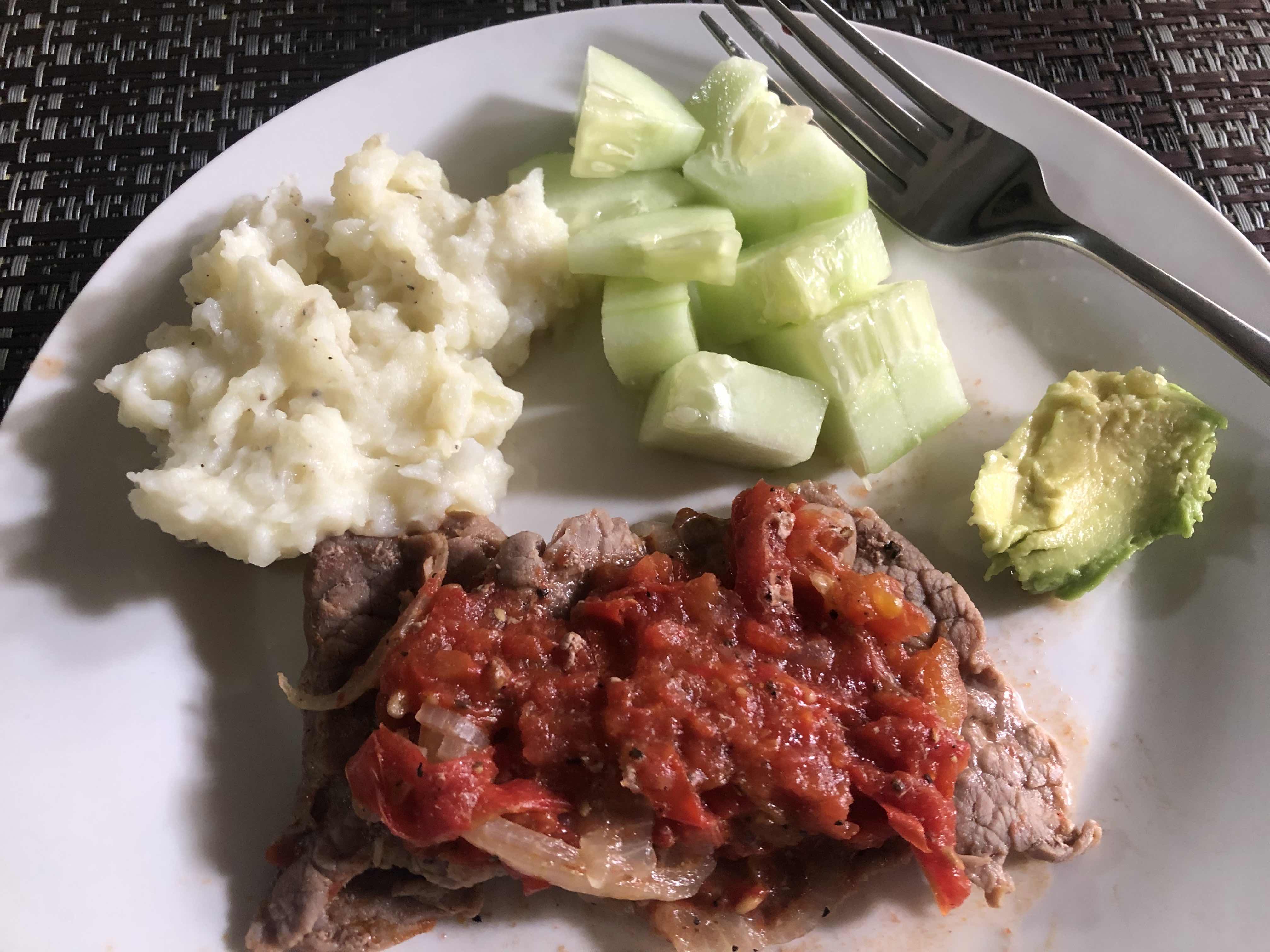 A balanced meal consisting of beef steak with tomato sauce, mashed potatoes, cucumber slices, and a small portion of avocado.