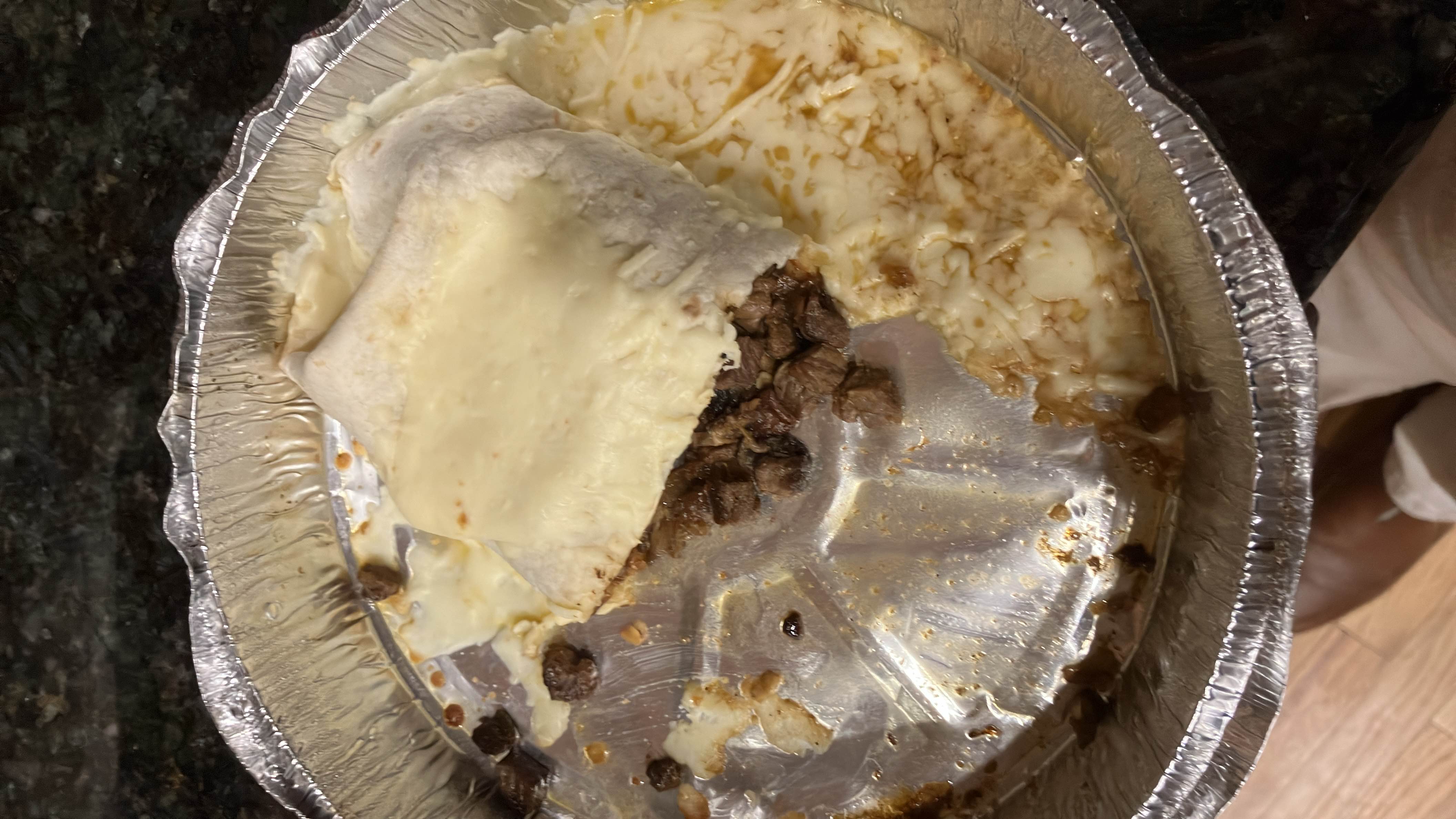 A partially eaten beef burrito covered in melted cheese, served in a foil container.