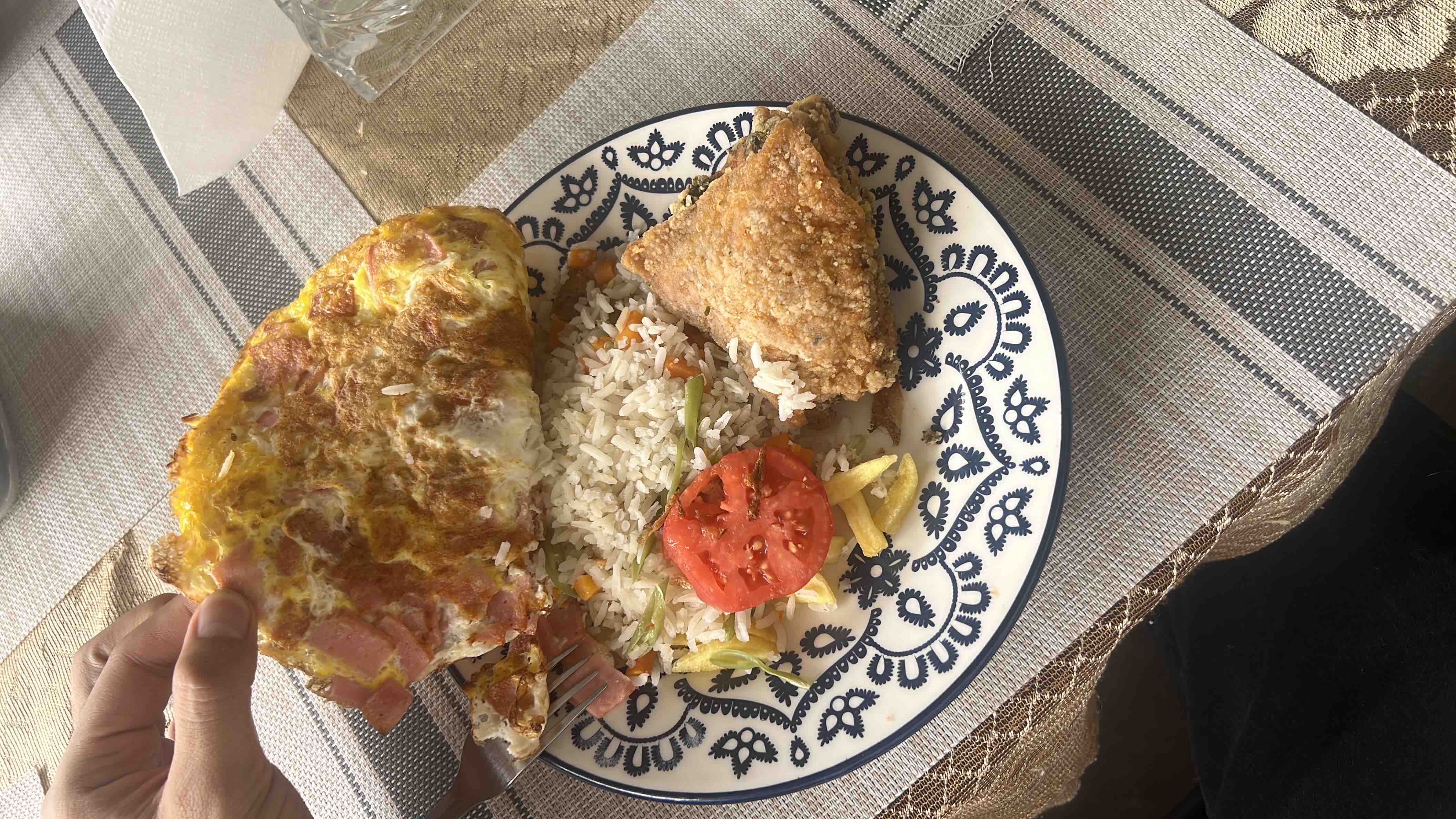 A plate containing an omelette with ham, white rice mixed with vegetables, a slice of tomato, and a piece of fried chicken.