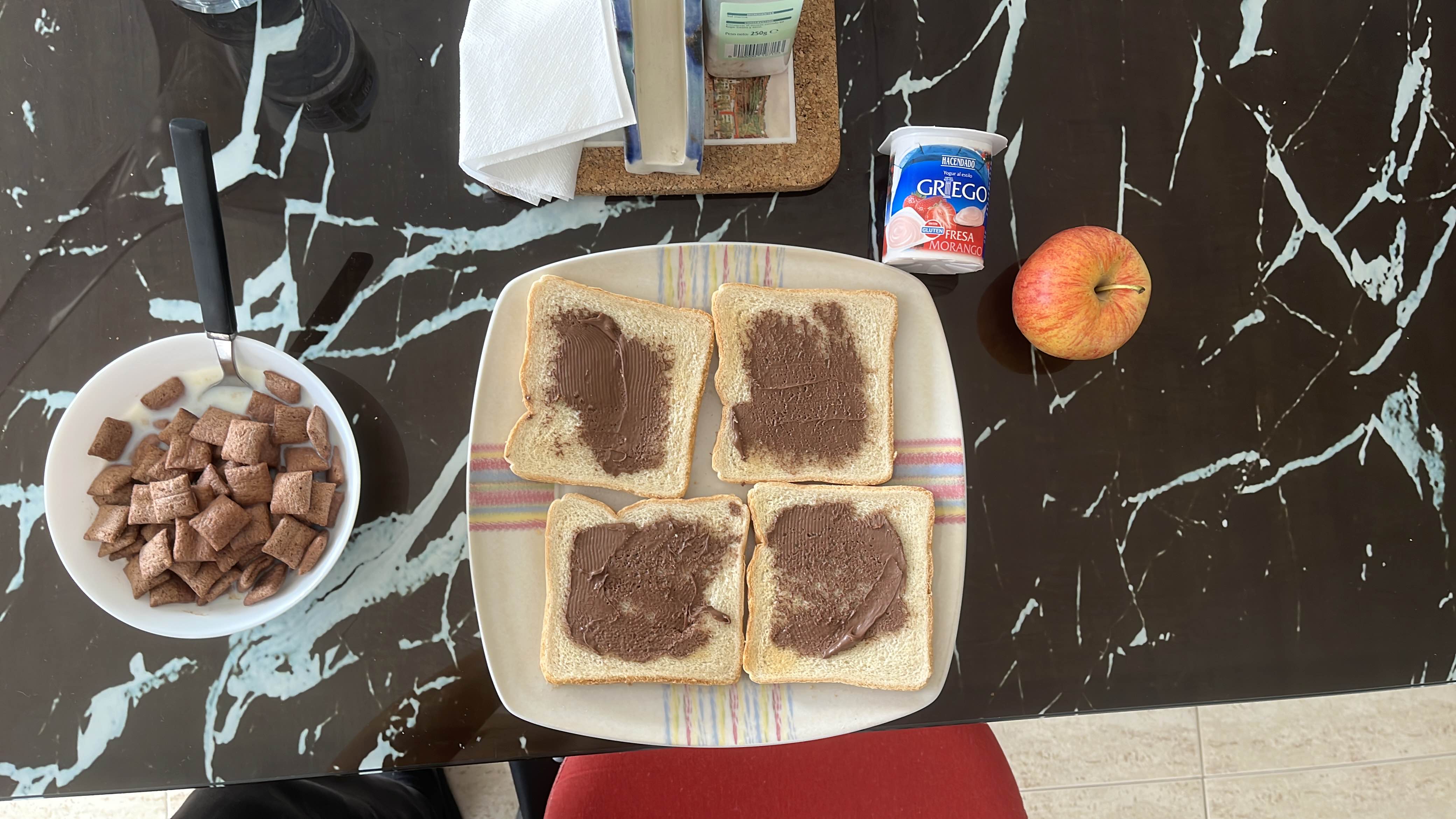 A breakfast meal consisting of bread with chocolate spread, cereal with milk, an apple, and strawberry yogurt.