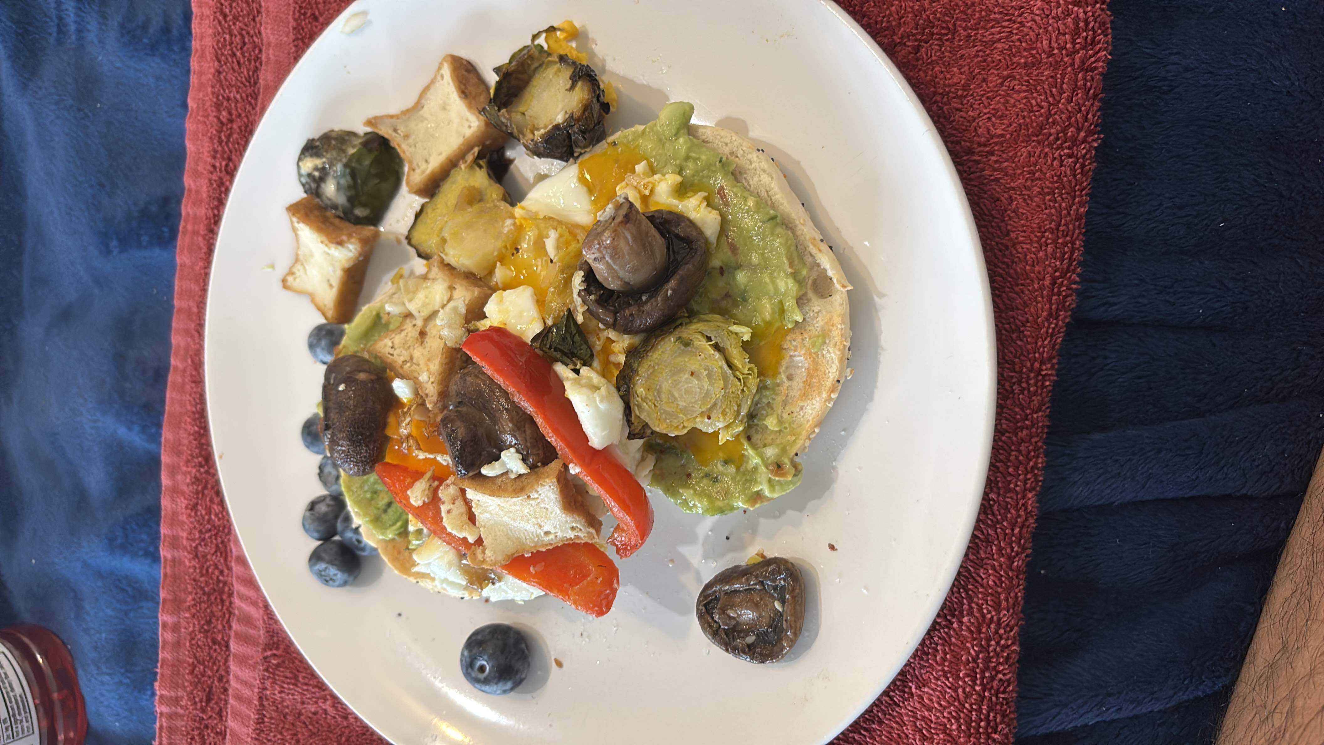 A nutritious plate featuring a bagel topped with avocado, egg, mushrooms, and roasted vegetables, accompanied by blueberries.