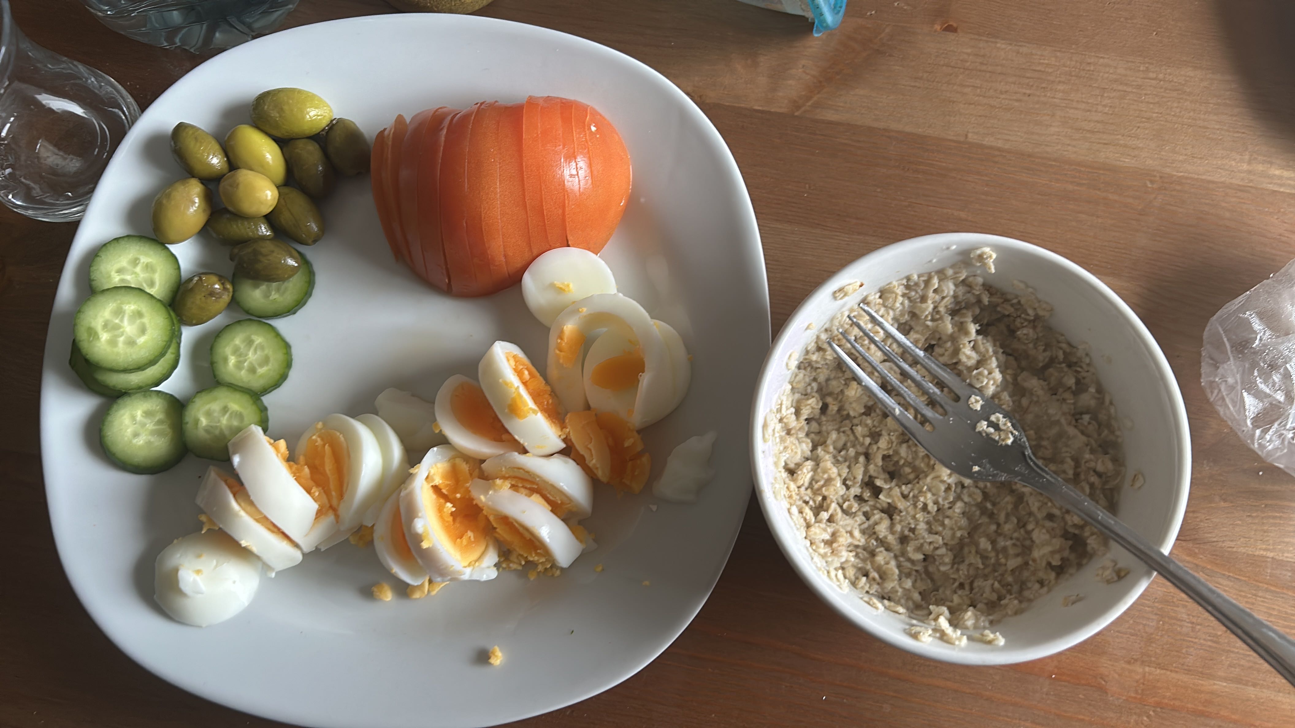A balanced meal consisting of boiled eggs, cucumber slices, olives, tomato slices, and a bowl of oatmeal.