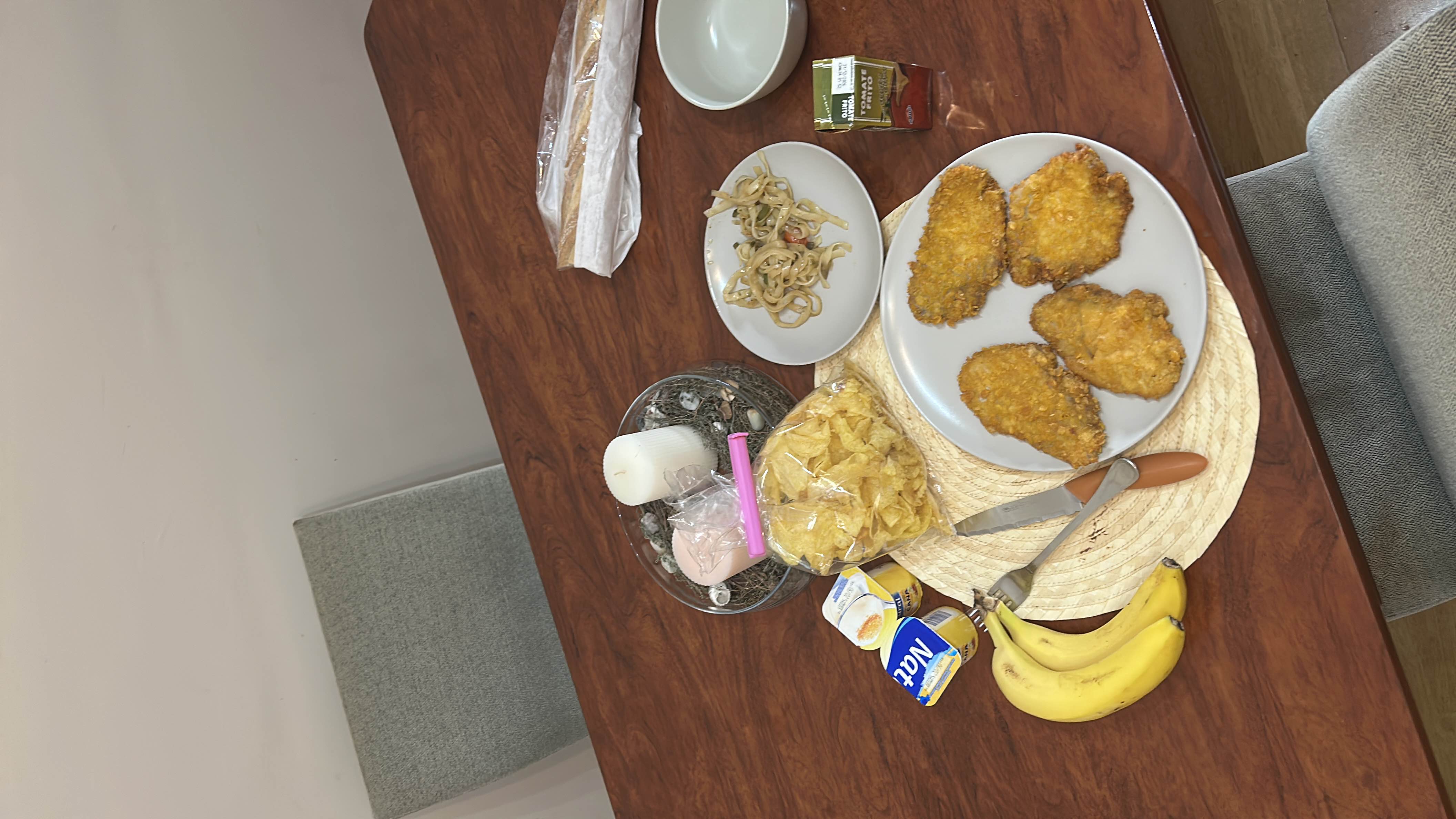 A meal consisting of fried chicken, pasta salad, bananas, potato chips, tomato juice, and natural yogurts, providing a balanced mix of protein, carbs, and fats.