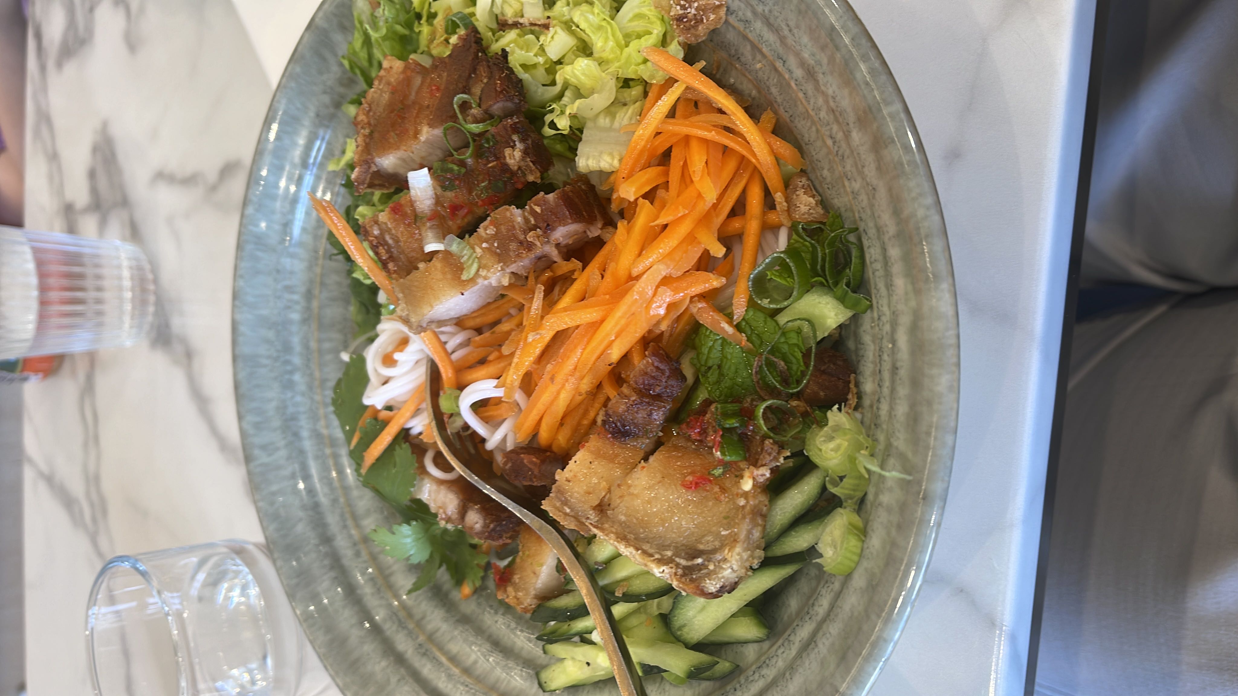 A balanced dish featuring crispy pork belly, rice noodles, and fresh vegetables like carrots, cucumber, and lettuce, offering a mix of protein, carbs, and fats.