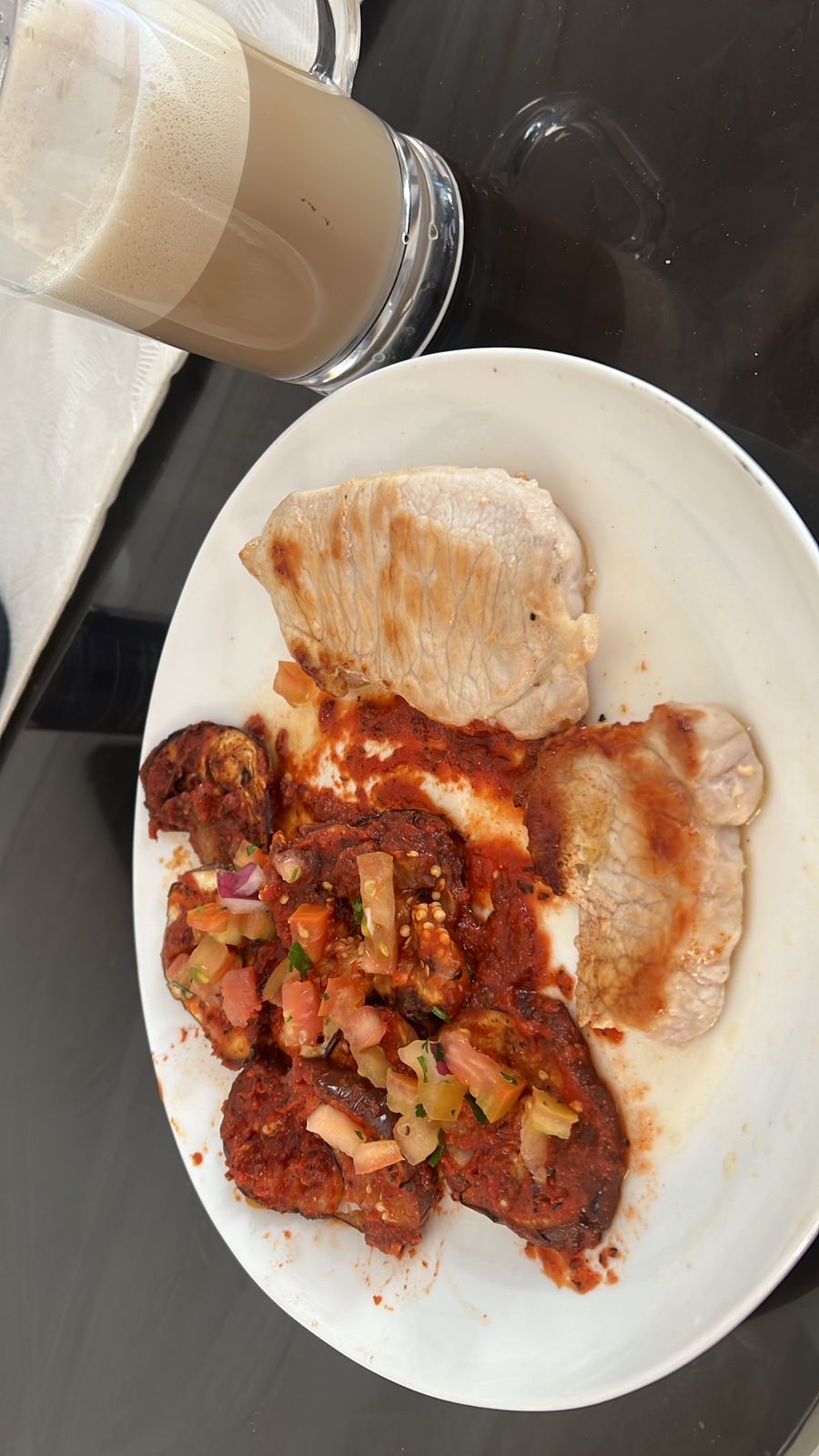A balanced meal consisting of grilled pork, eggplant in tomato sauce, vegetable salsa, and a glass of coffee with milk.