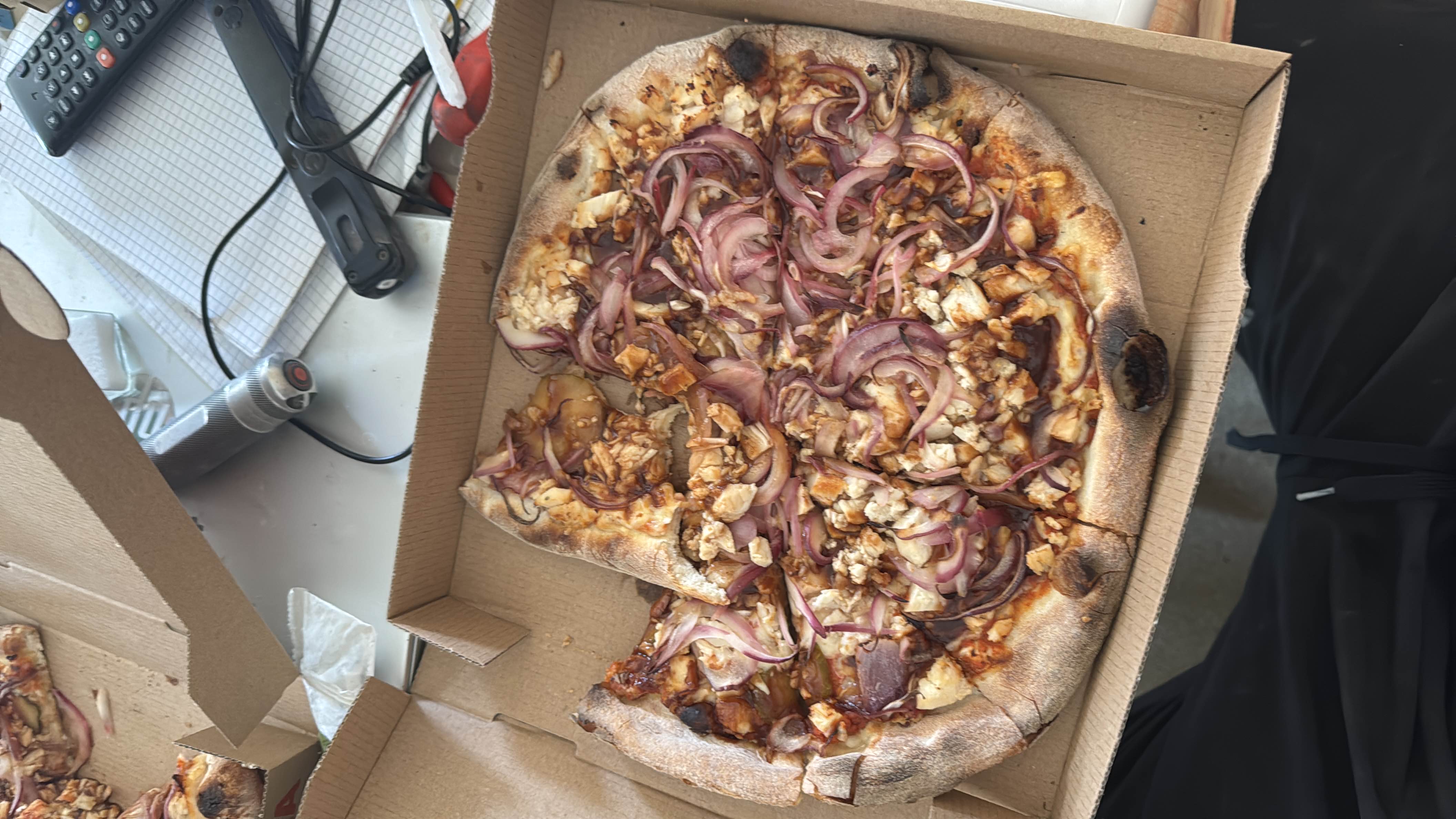 A barbecue chicken pizza topped with red onions, cheese, and barbecue sauce, served in a cardboard box.