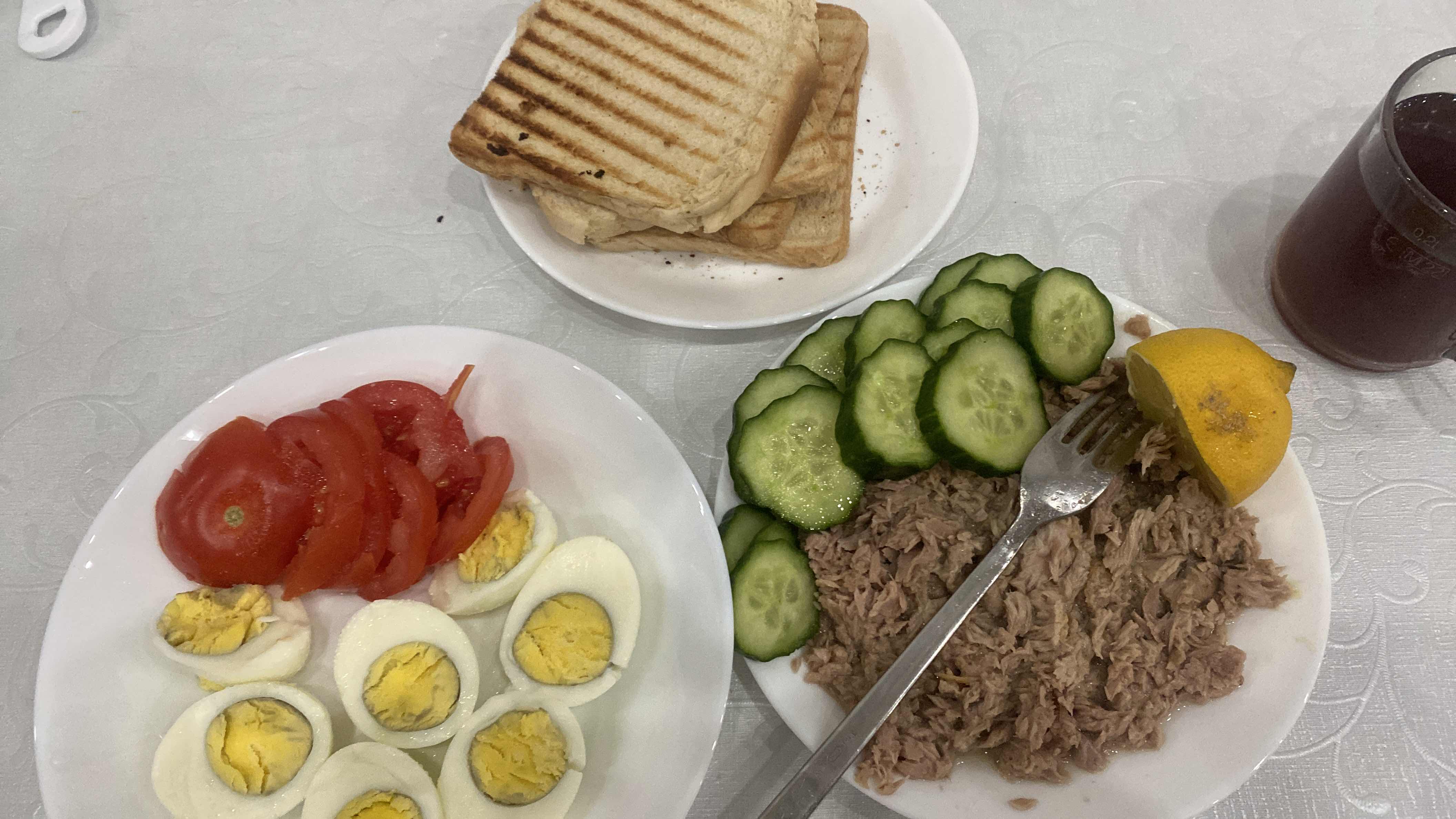 A balanced meal consisting of boiled eggs, sliced tomatoes, cucumbers, tuna, grilled bread, and a lemon wedge, served with a beverage.
