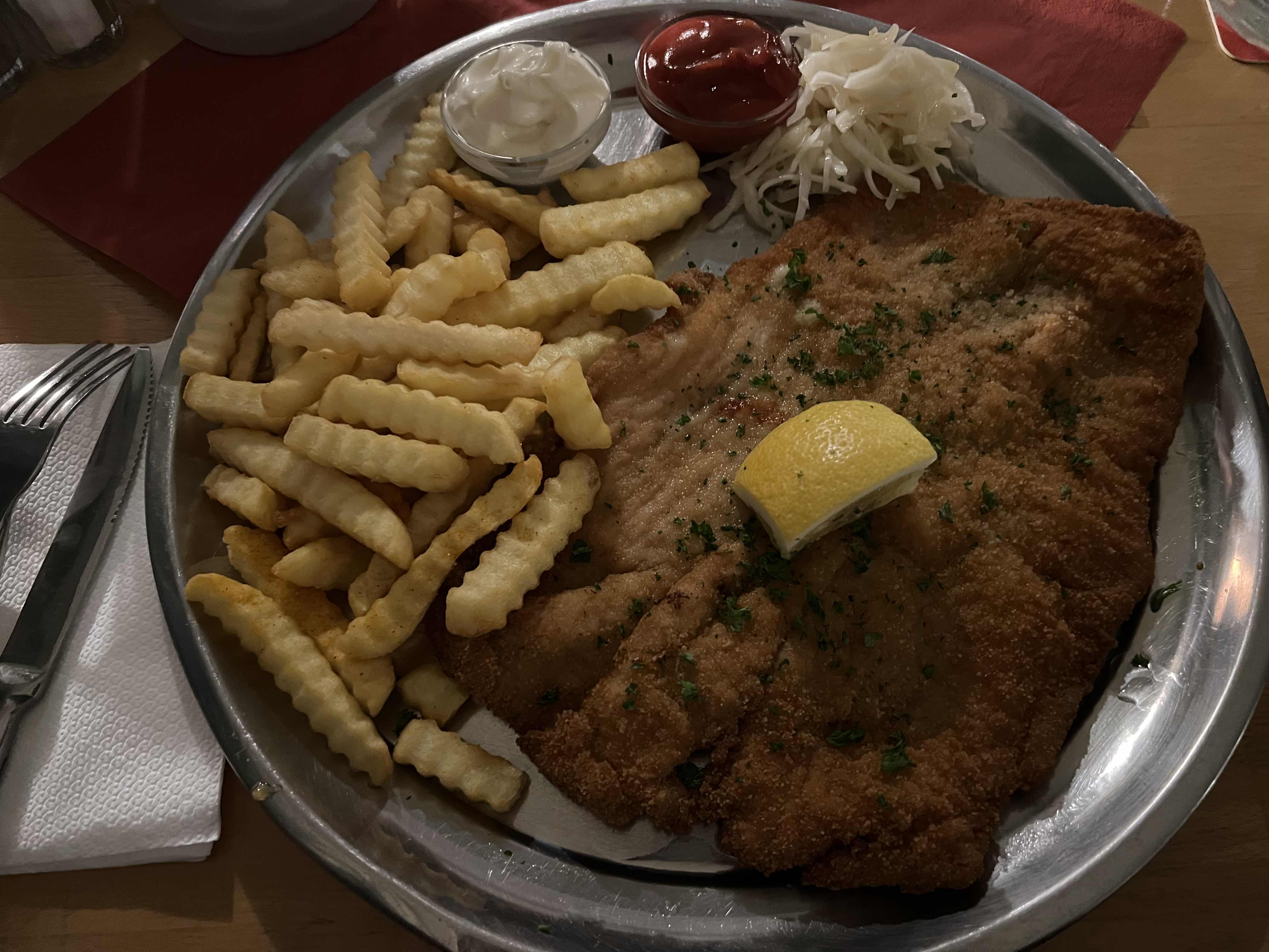A plate of breaded schnitzel served with french fries, mayonnaise, ketchup, a lemon wedge, and a side of shredded cabbage.