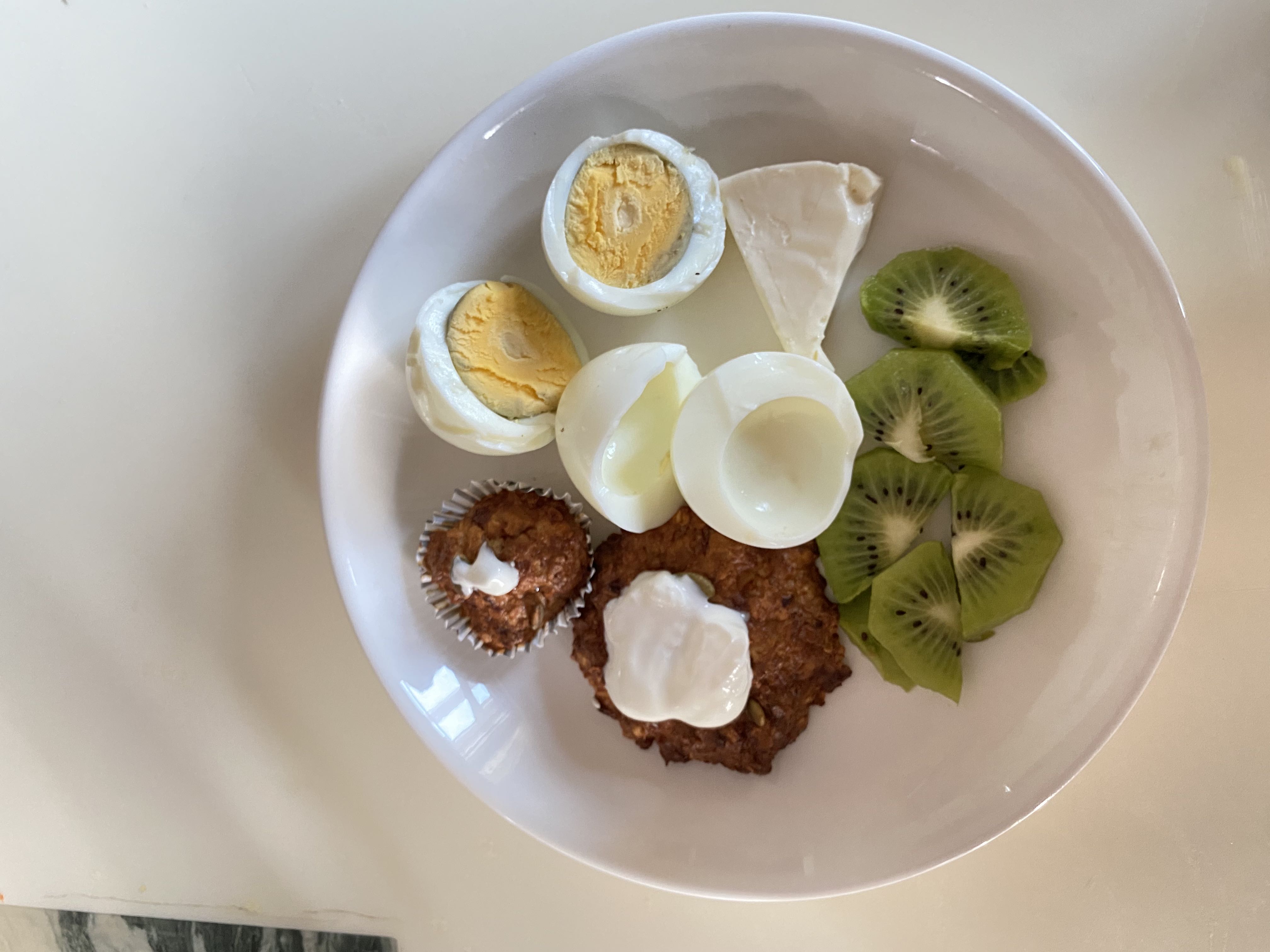 A balanced plate containing boiled eggs, kiwi slices, brie cheese, an oat pancake with yogurt, and a mini muffin.
