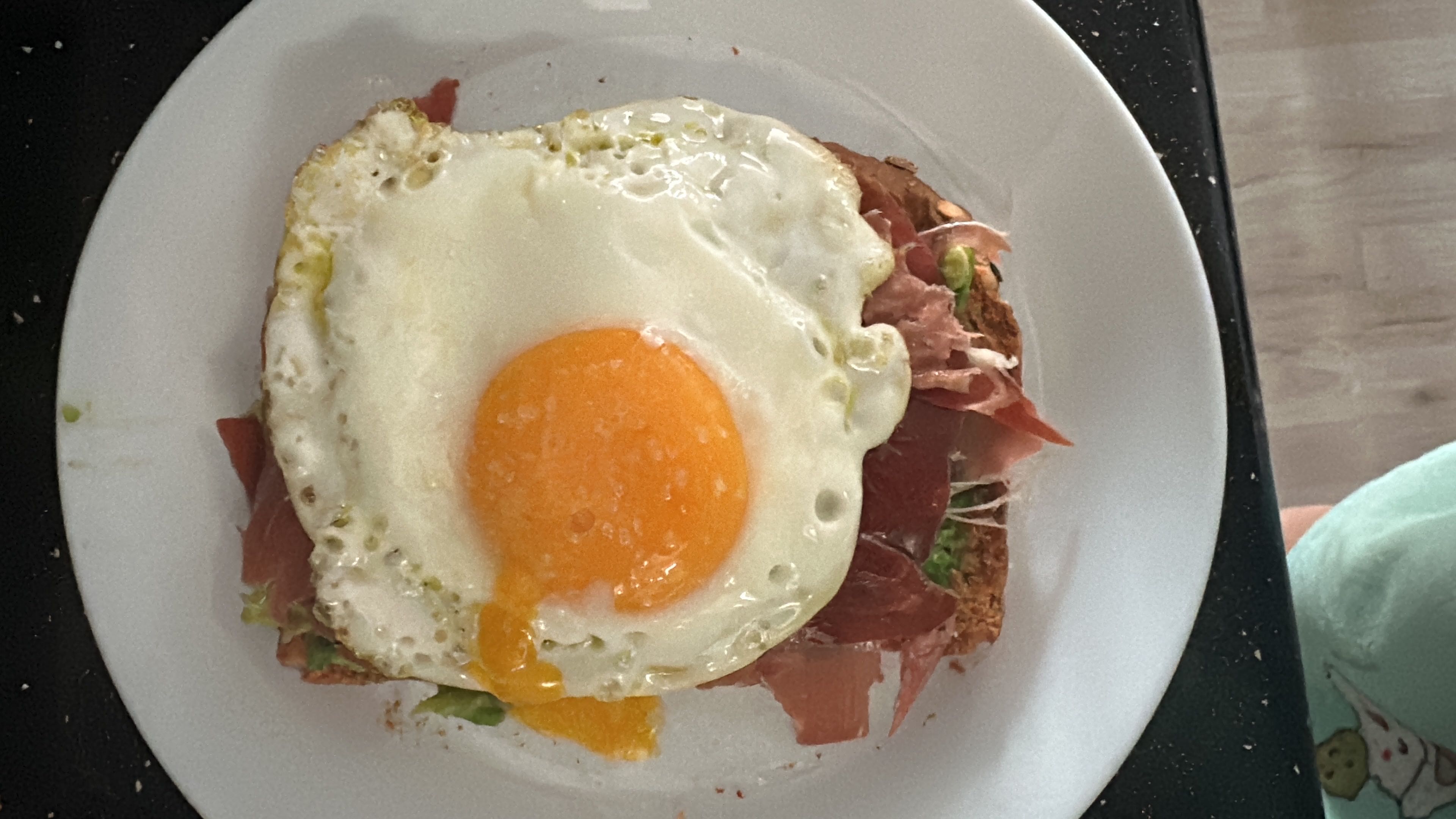 A nutritious open-faced sandwich featuring a fried egg, avocado, prosciutto, and toast.