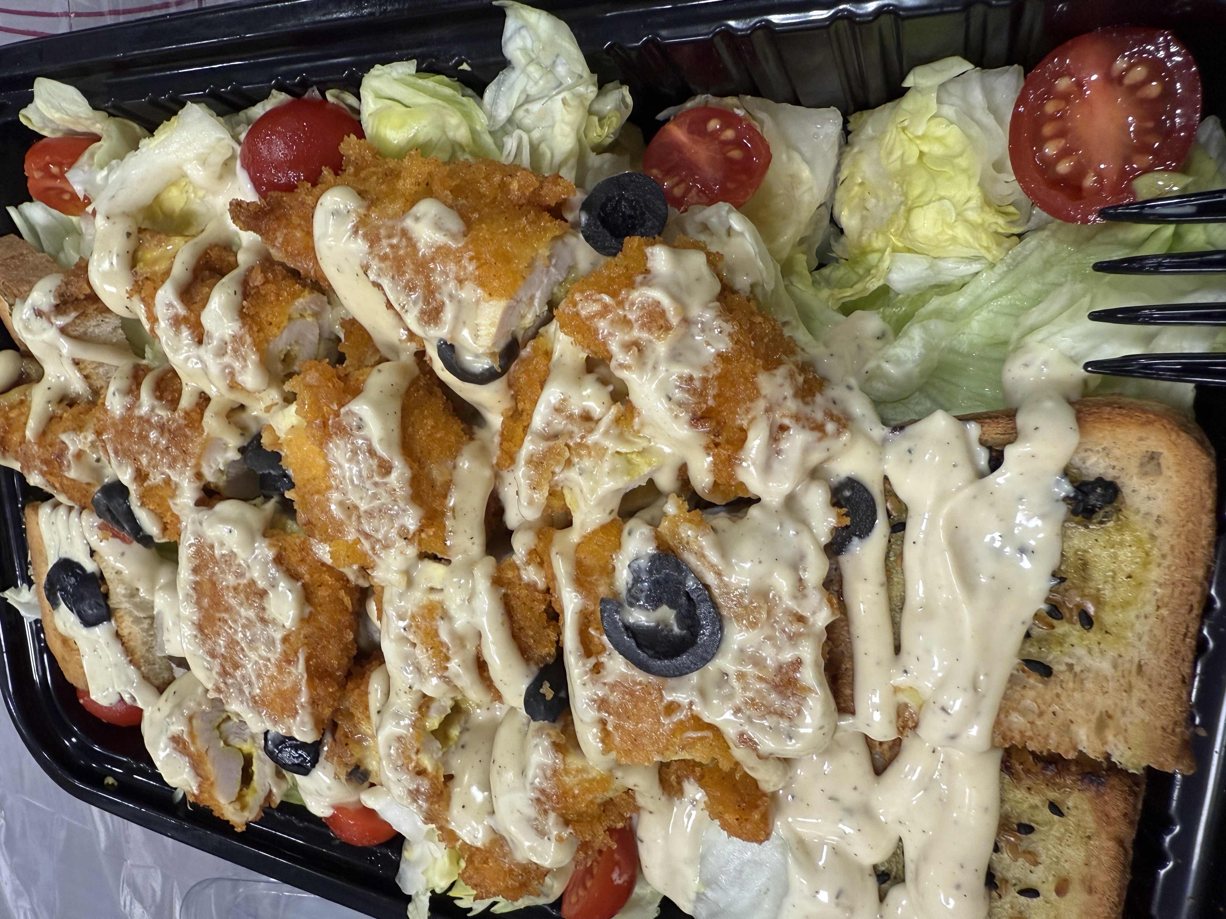 A dish consisting of breaded chicken, lettuce, cherry tomatoes, black olives, garlic bread, and creamy dressing.