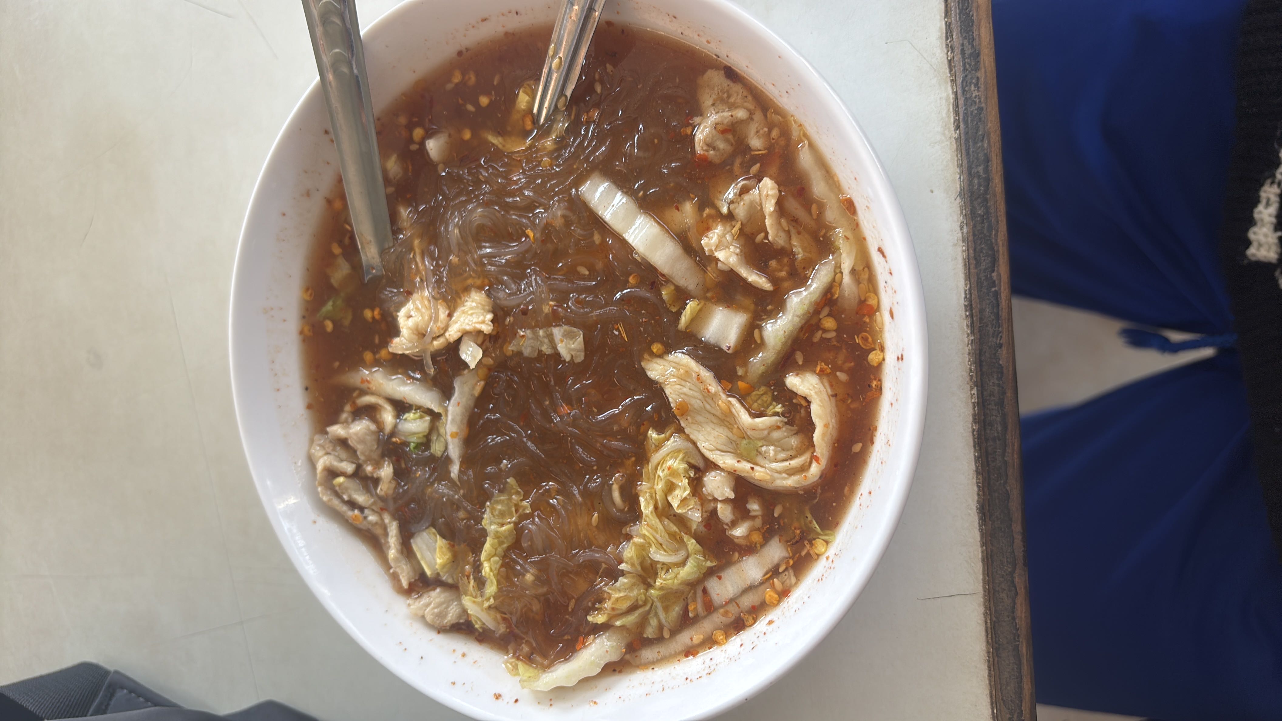 A bowl of spicy glass noodle soup with chicken slices, cabbage, and a flavorful broth.