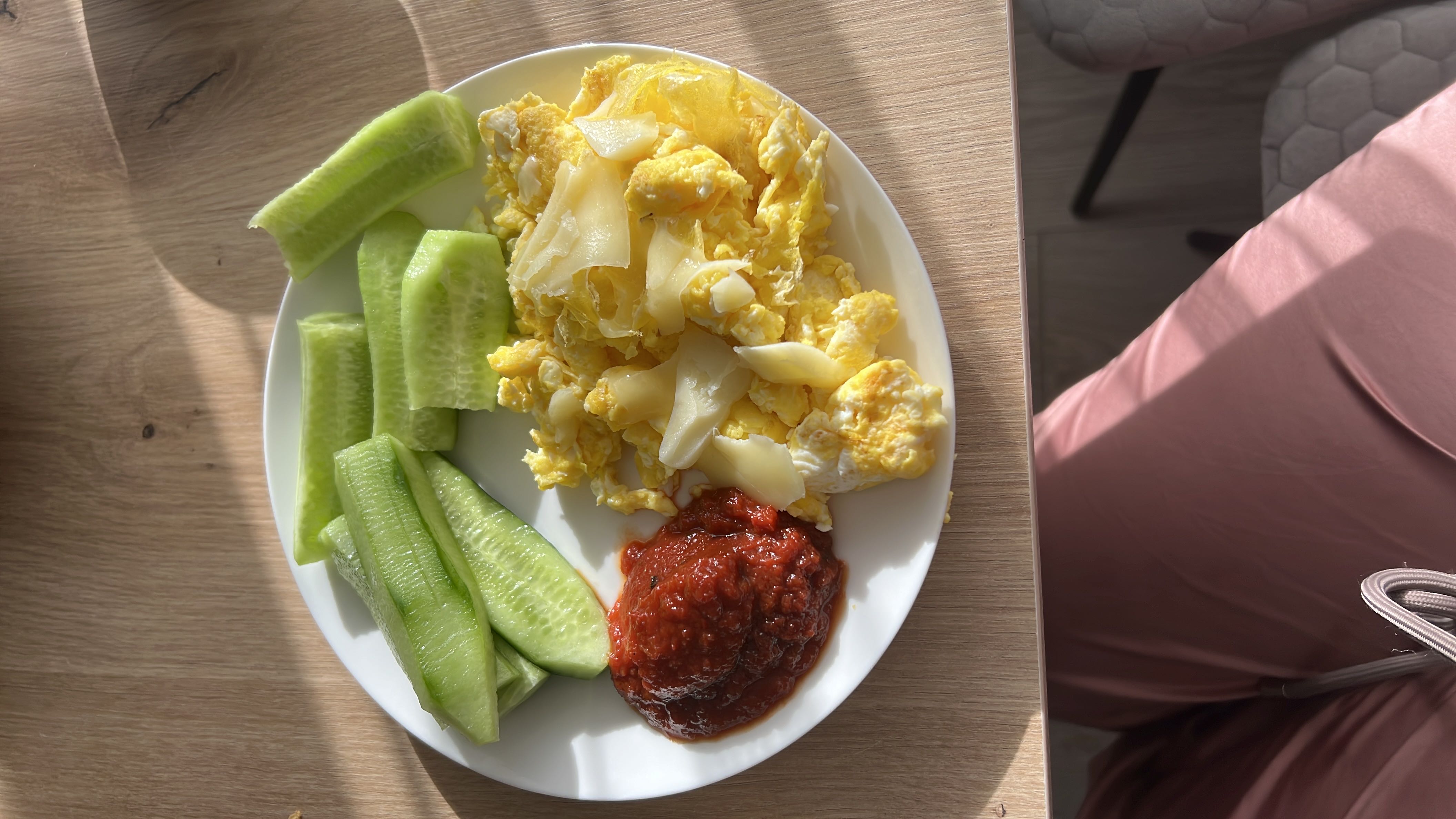 A balanced plate with scrambled eggs topped with cheese, fresh cucumber slices, and a serving of tomato sauce.