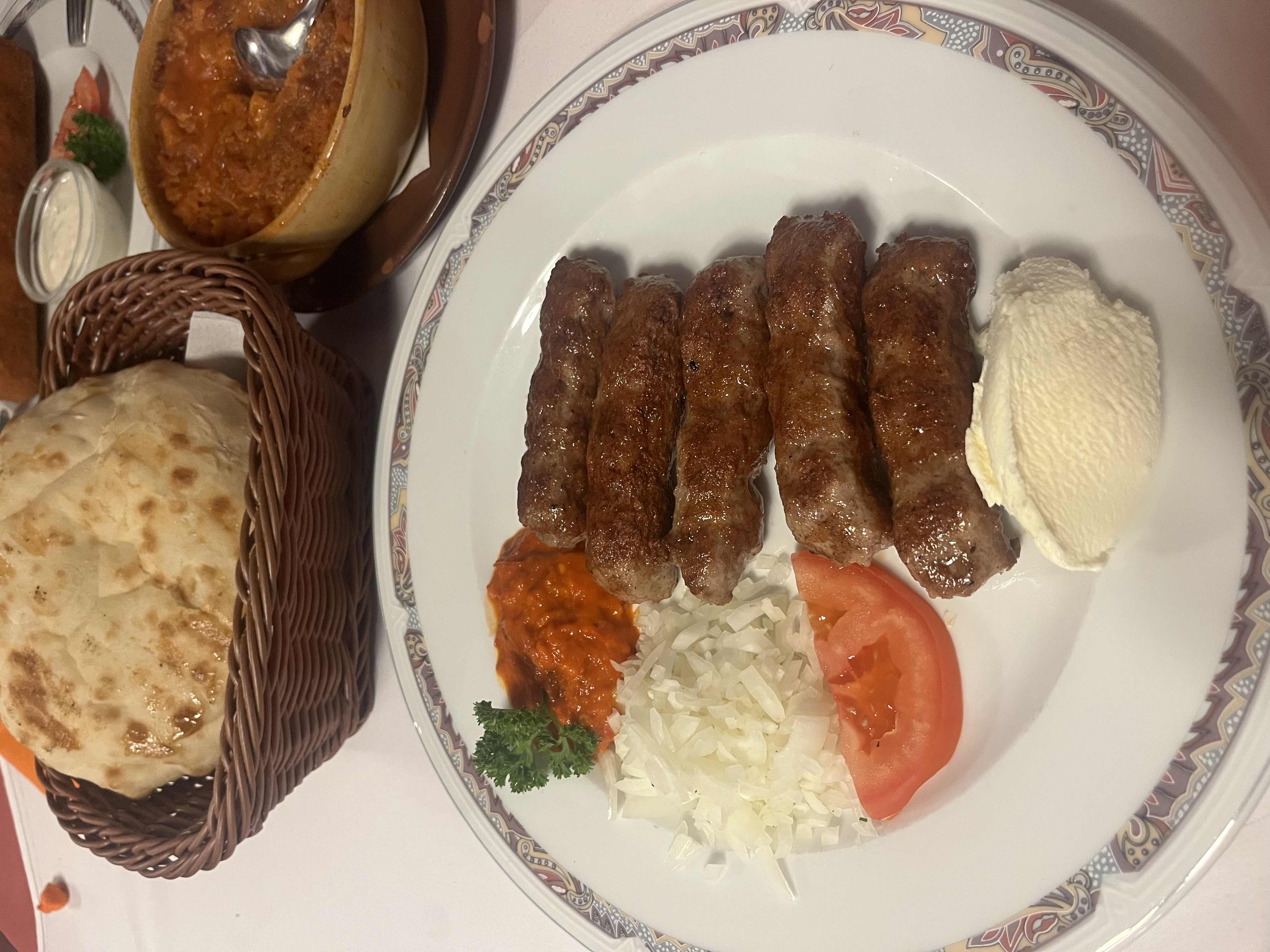 A traditional meal featuring grilled sausages, flatbread, ajvar, onions, tomato, cream, and a side stew.