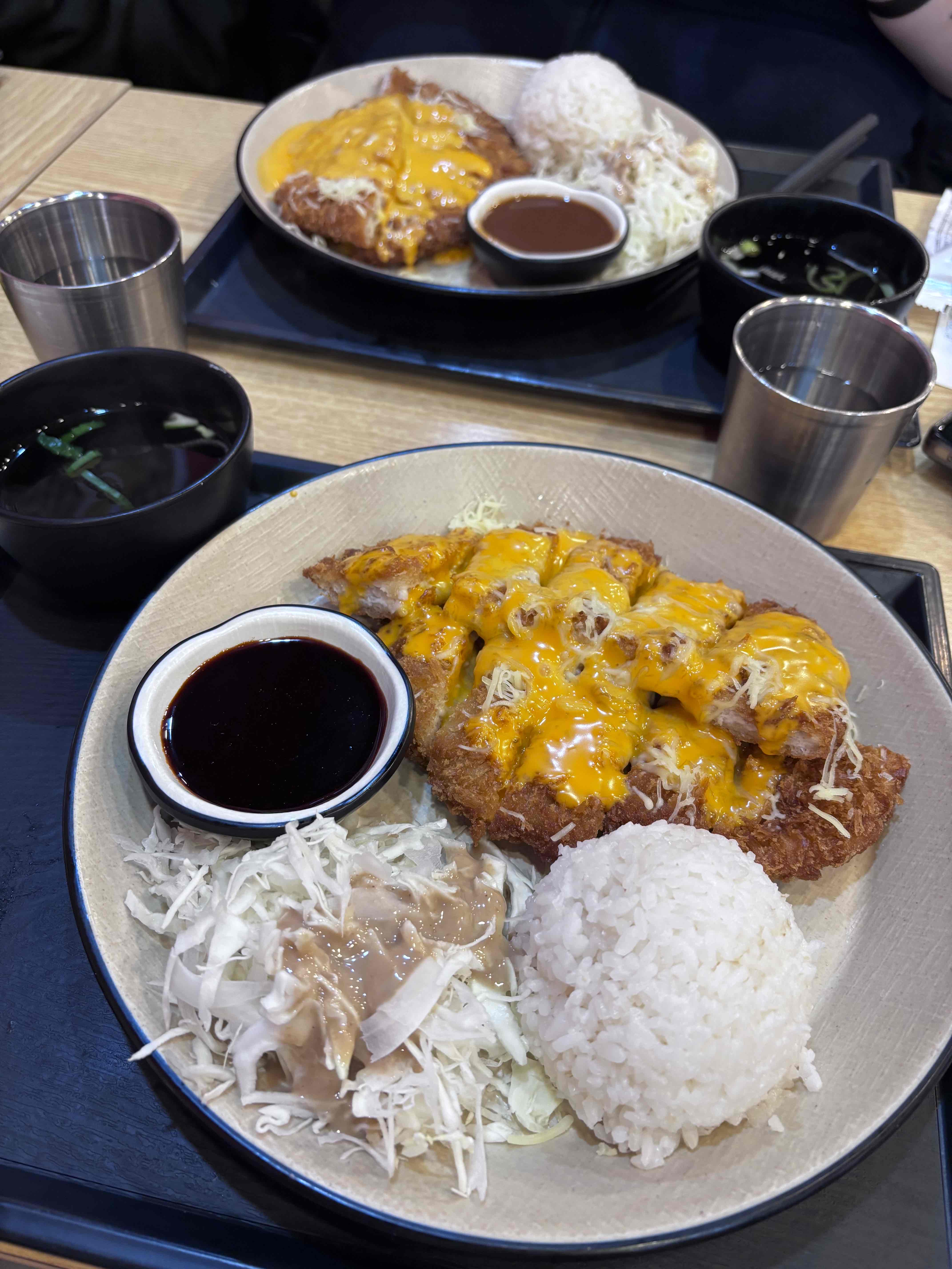 A meal consisting of fried pork cutlet topped with cheese, served with steamed rice, shredded cabbage, and dipping sauce.