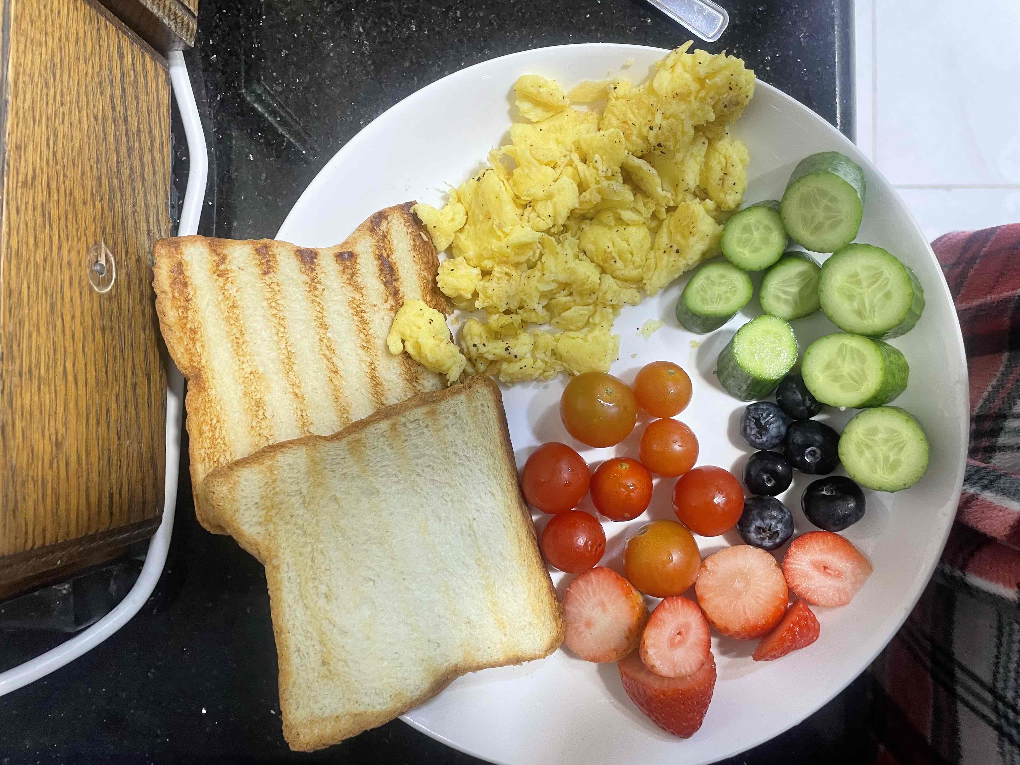 A balanced breakfast plate containing scrambled eggs, toast, cherry tomatoes, strawberries, blueberries, and cucumber slices.
