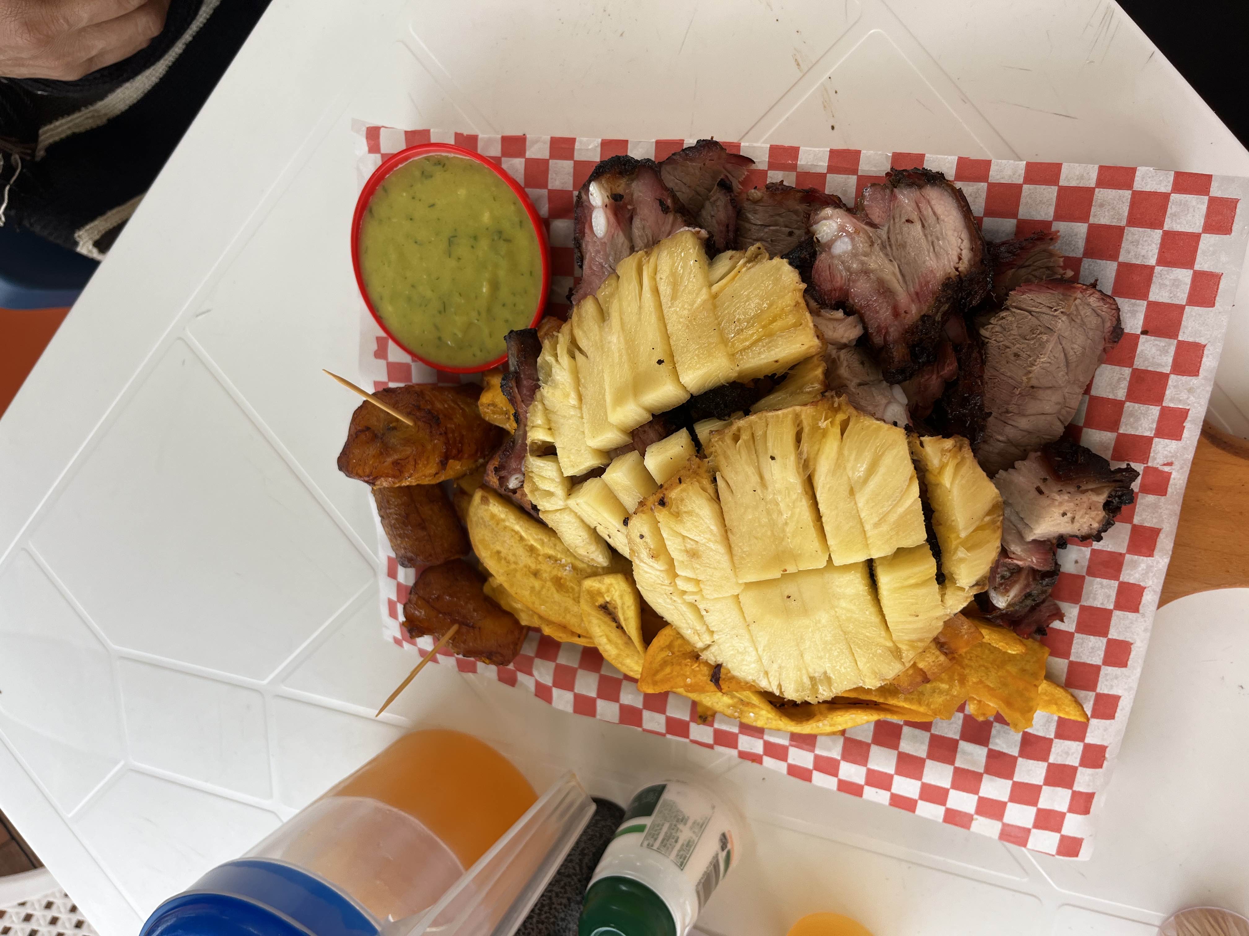 A dish featuring grilled meat, fresh pineapple slices, fried plantains, chips, and a green dipping sauce.