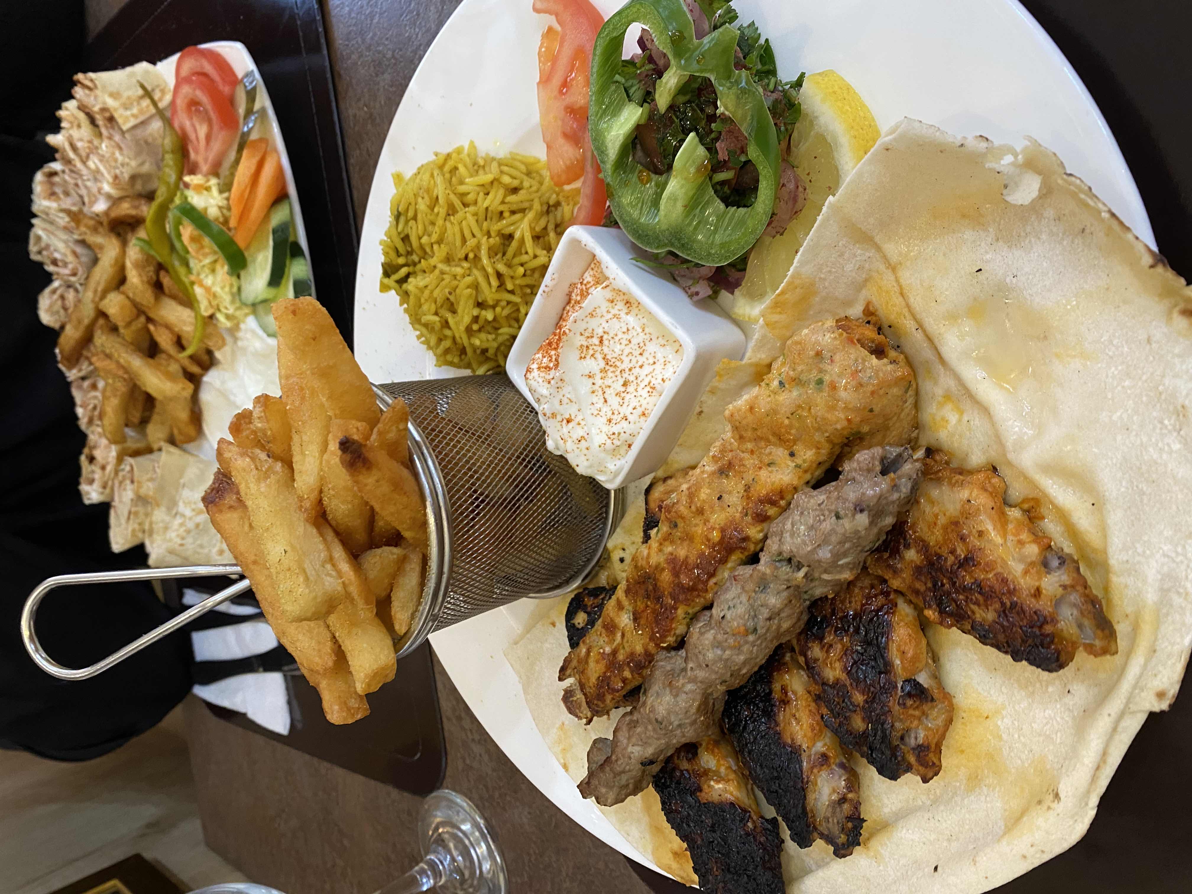 A mixed platter featuring grilled chicken, beef kebab, French fries, rice, and a side salad with yogurt dip.