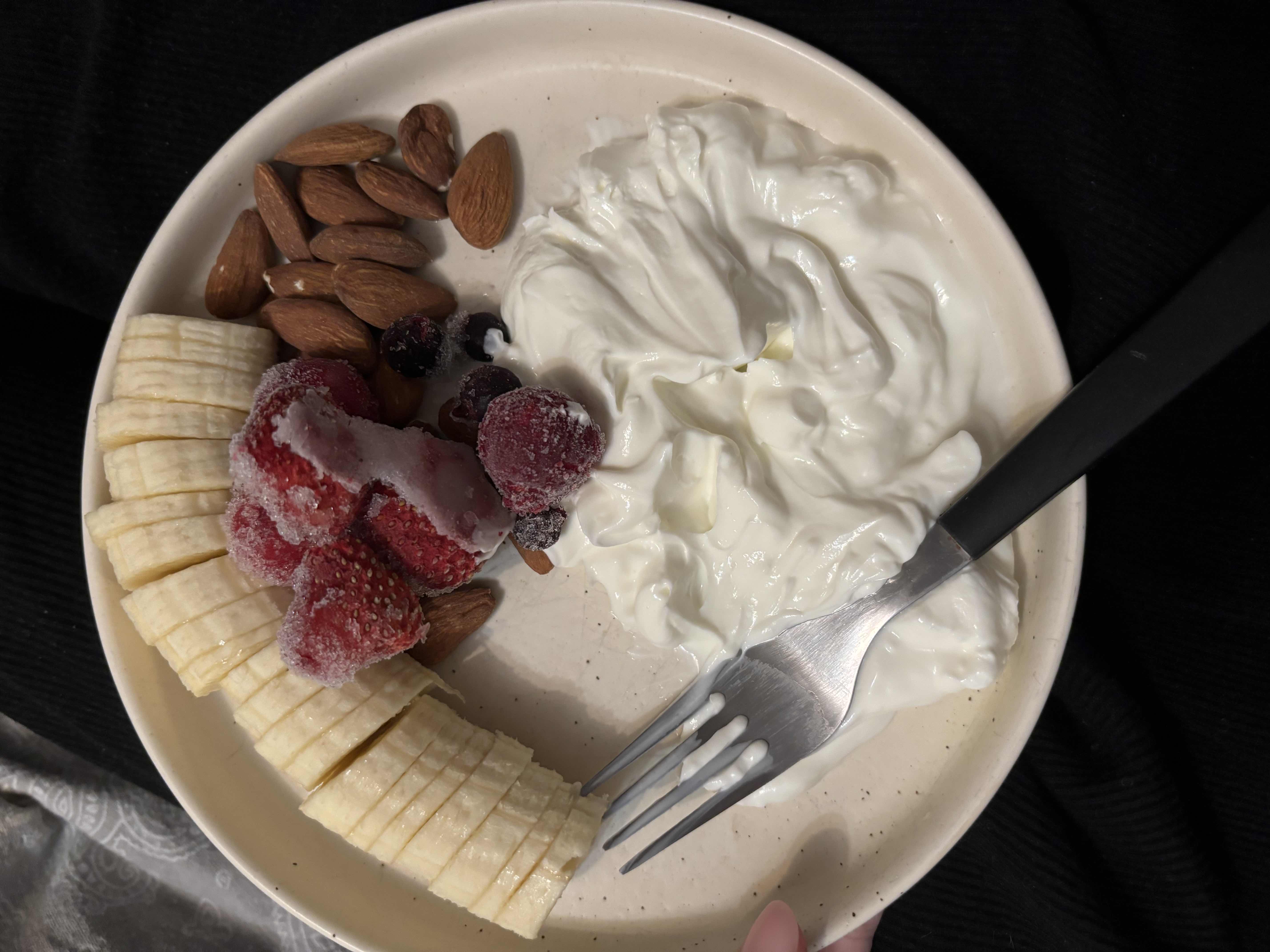 A healthy snack consisting of sliced banana, frozen berries, almonds, and Greek yogurt, providing a balanced mix of protein, carbs, and fats.