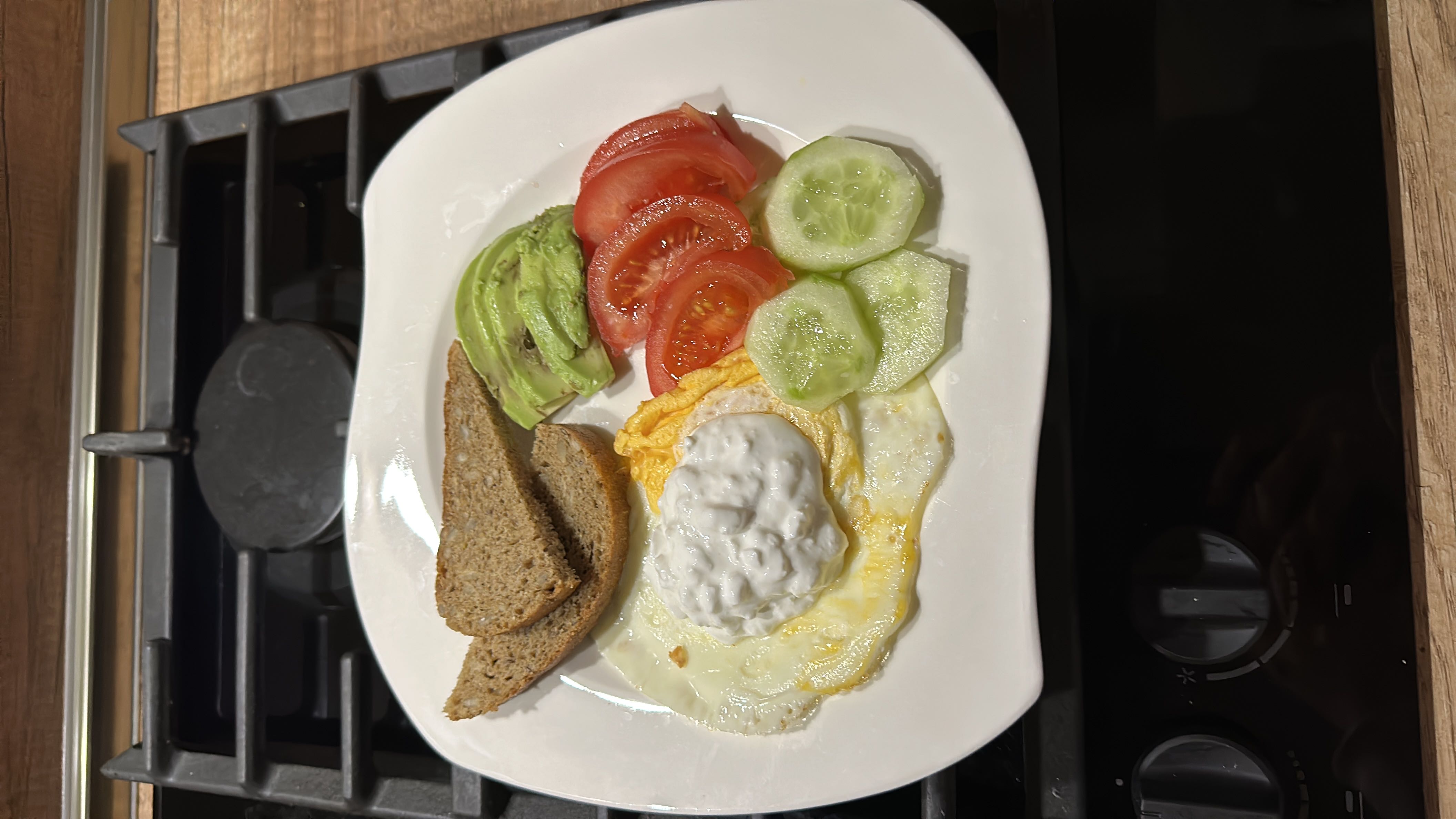 A balanced meal consisting of fried egg, avocado slices, tomato slices, cucumber slices, whole grain bread, and a dollop of cottage cheese.
