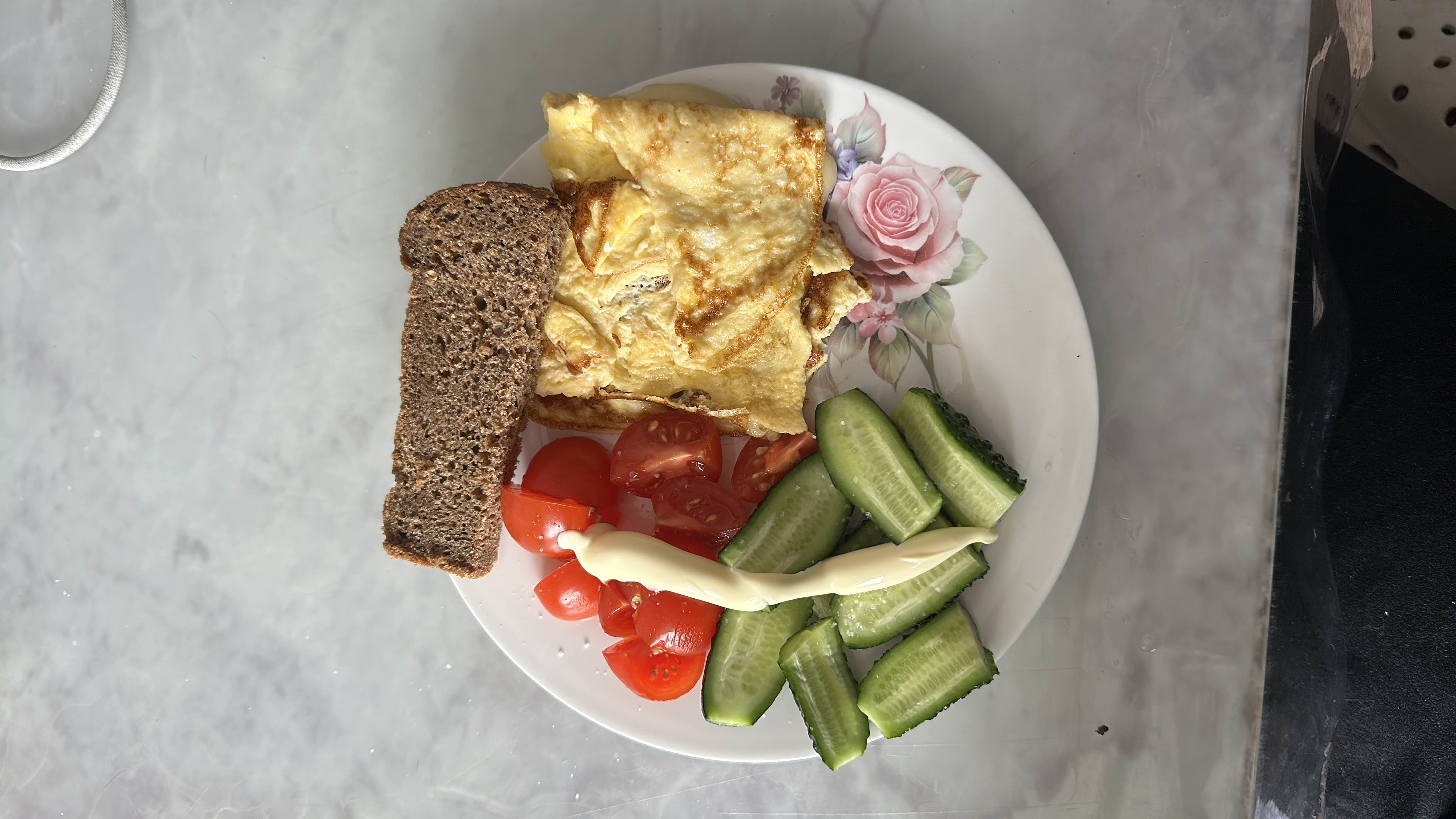 A balanced meal consisting of an egg omelette, fresh cucumber and tomato slices, rye bread, and a drizzle of mayonnaise.