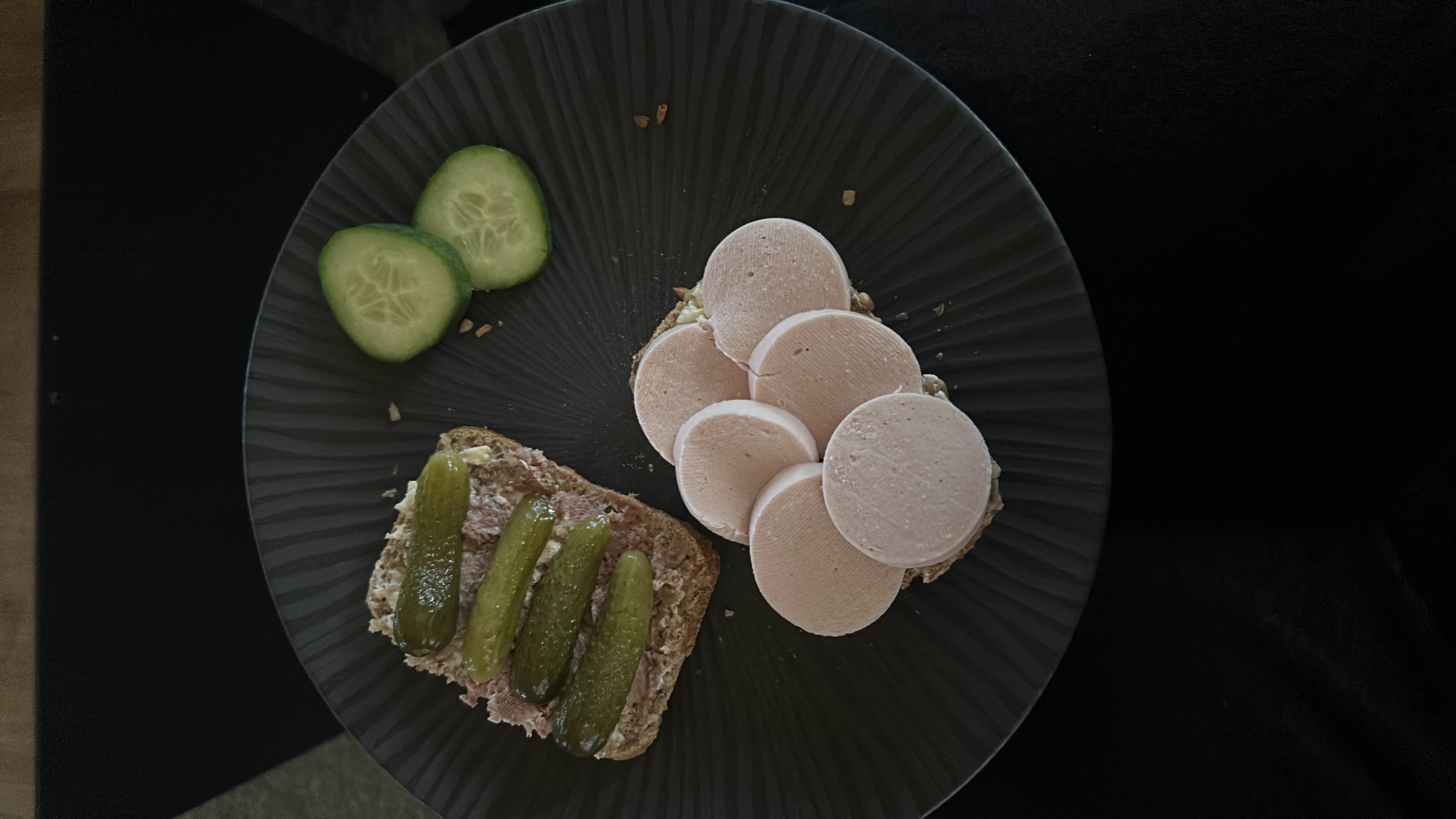 A plate with an open-faced sandwich topped with liver pate and pickles, another sandwich with bologna slices, and cucumber slices on the side.