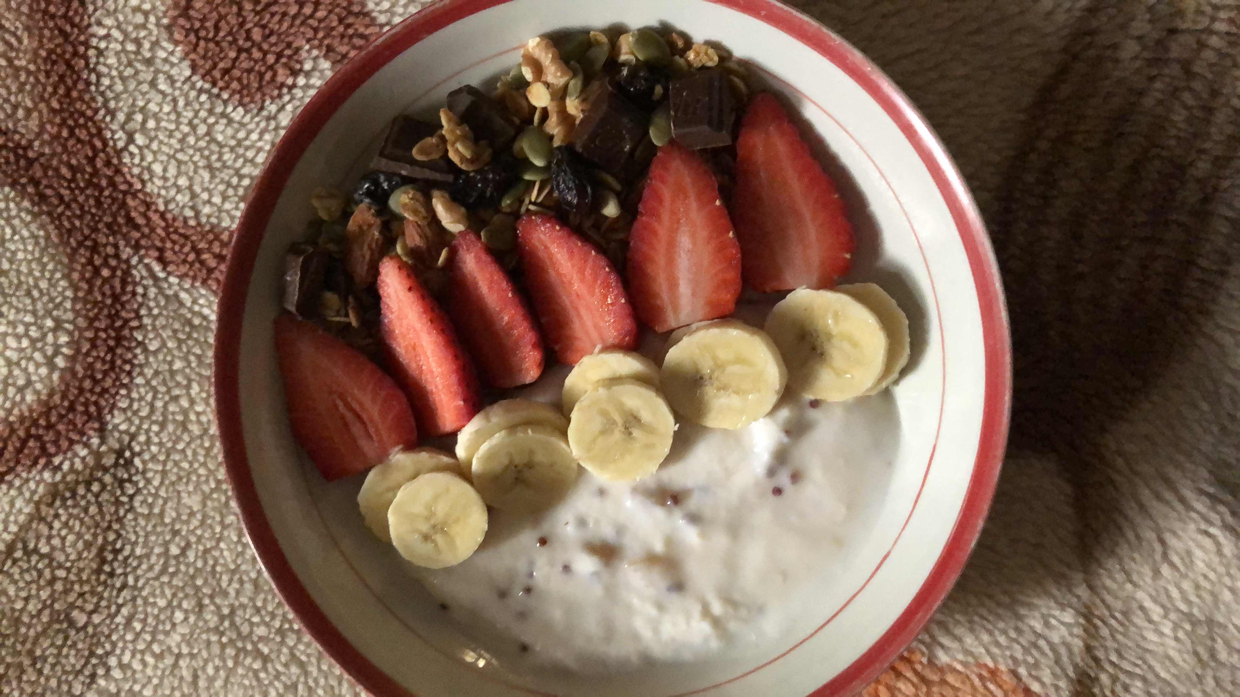 A nutritious bowl containing yogurt, banana slices, strawberries, granola, and dark chocolate, offering a balanced mix of protein, carbs, and fats.