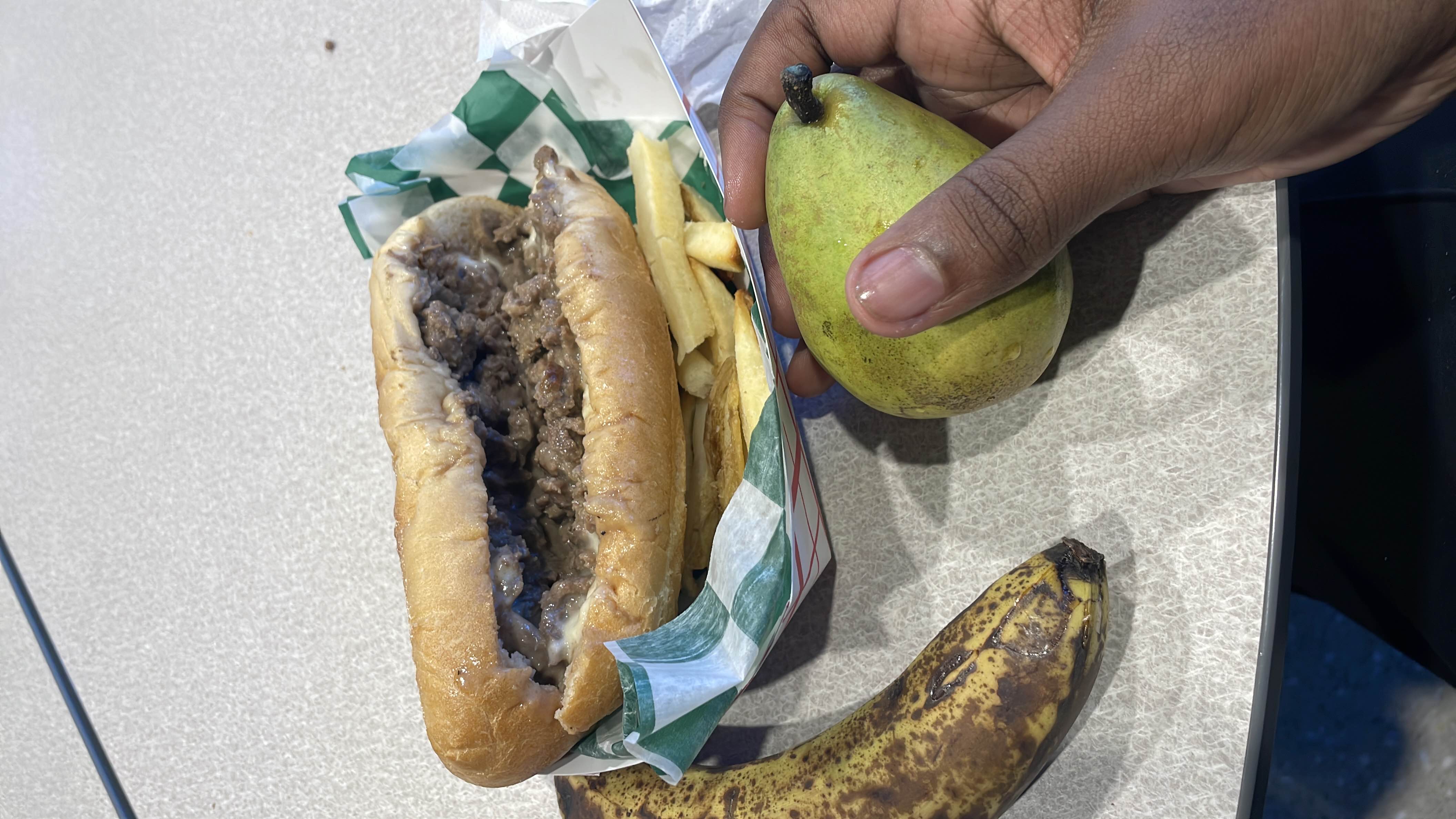 A meal consisting of a cheesesteak sandwich, french fries, a ripe banana, and a pear.